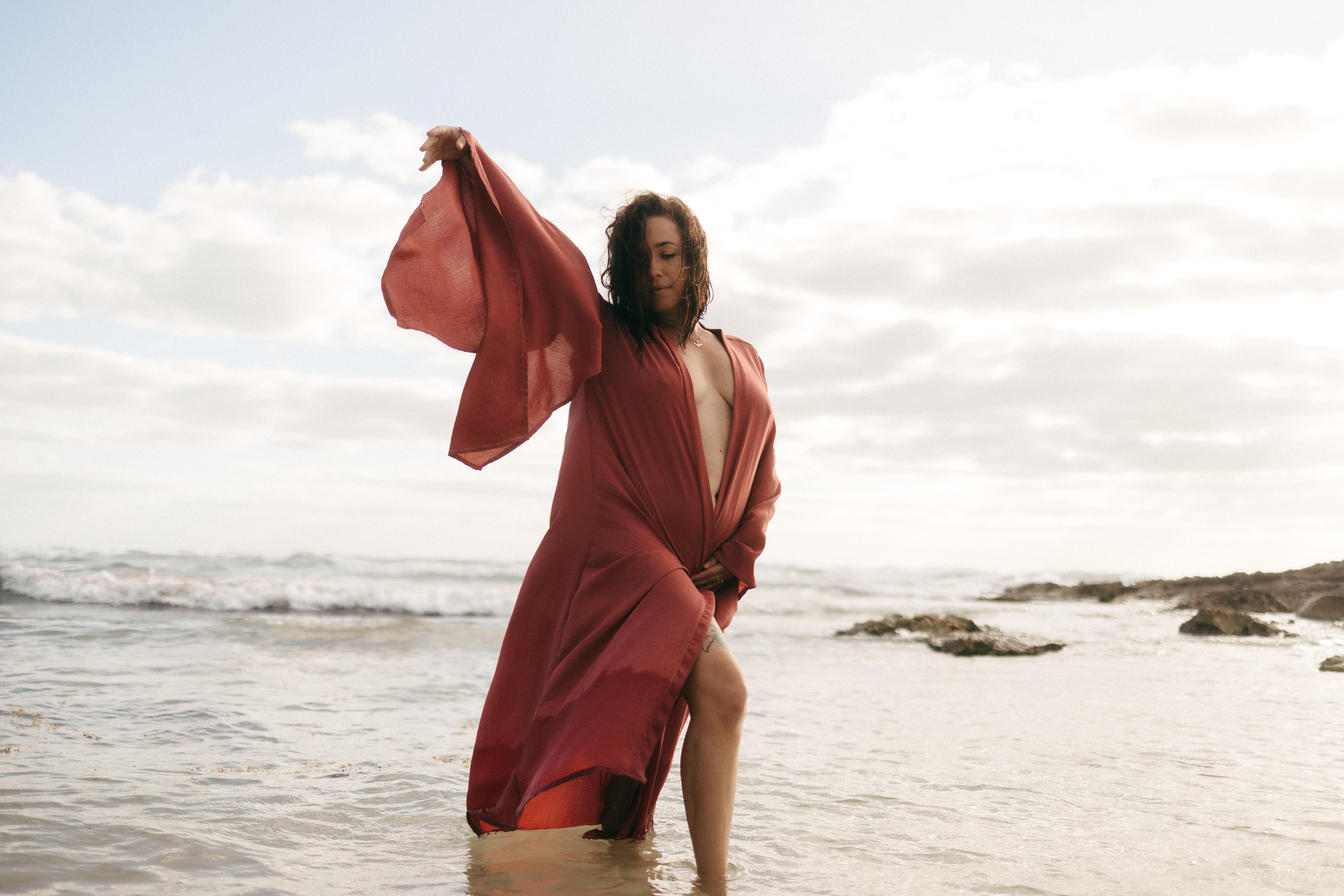 Woman in a red dress standing on a beach with ocean and cloudy sky in the background.