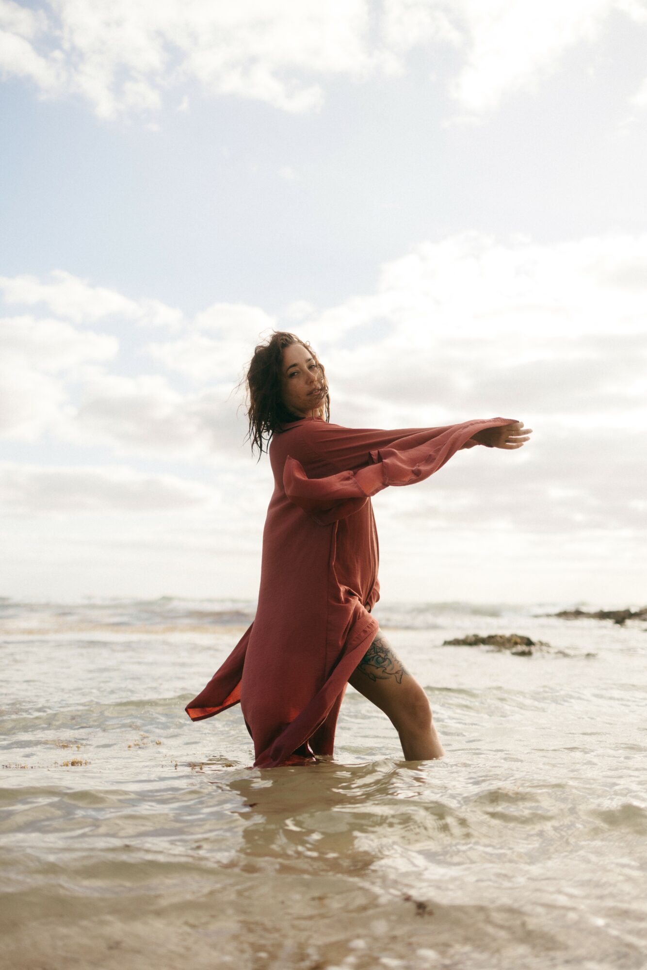 Woman standing in shallow water at the beach, wearing a long red dress, with clouds in the sky behind her.