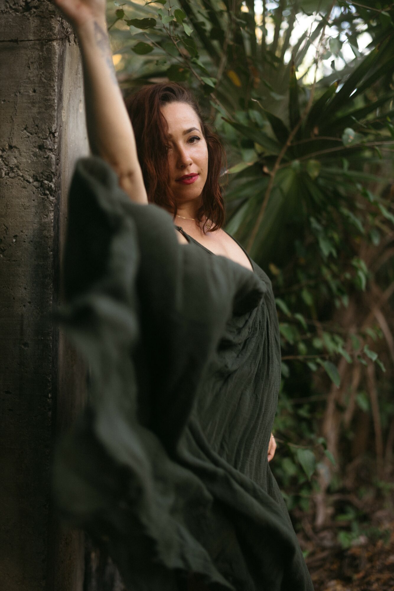 Woman with shoulder-length hair leaning against a tree in a lush outdoor setting, wearing a dark dress.
