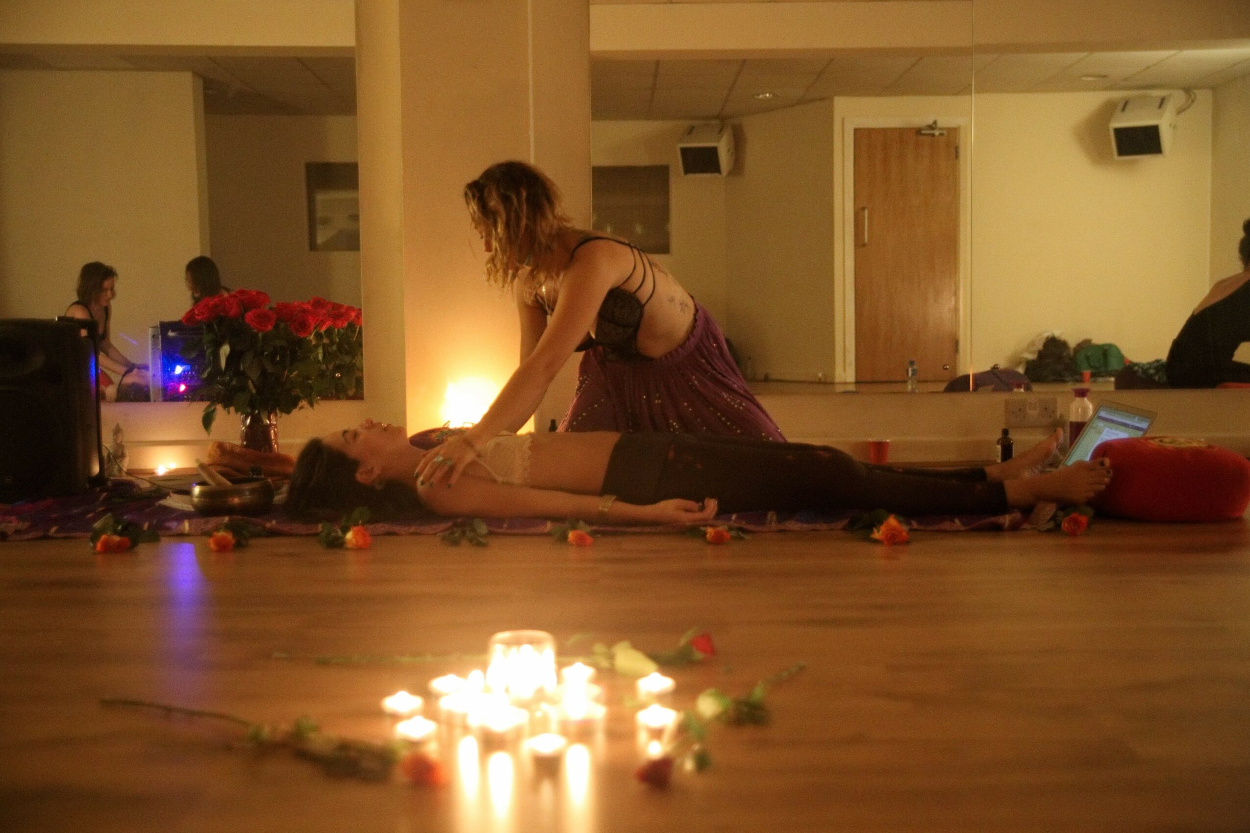 Person lying on the floor with another person kneeling beside them, candles and flowers on the floor, dim lighting.
