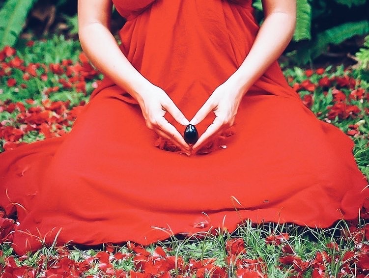 Person in a red dress sitting cross-legged outdoors, hands forming a heart shape, surrounded by red flowers and green foliage.