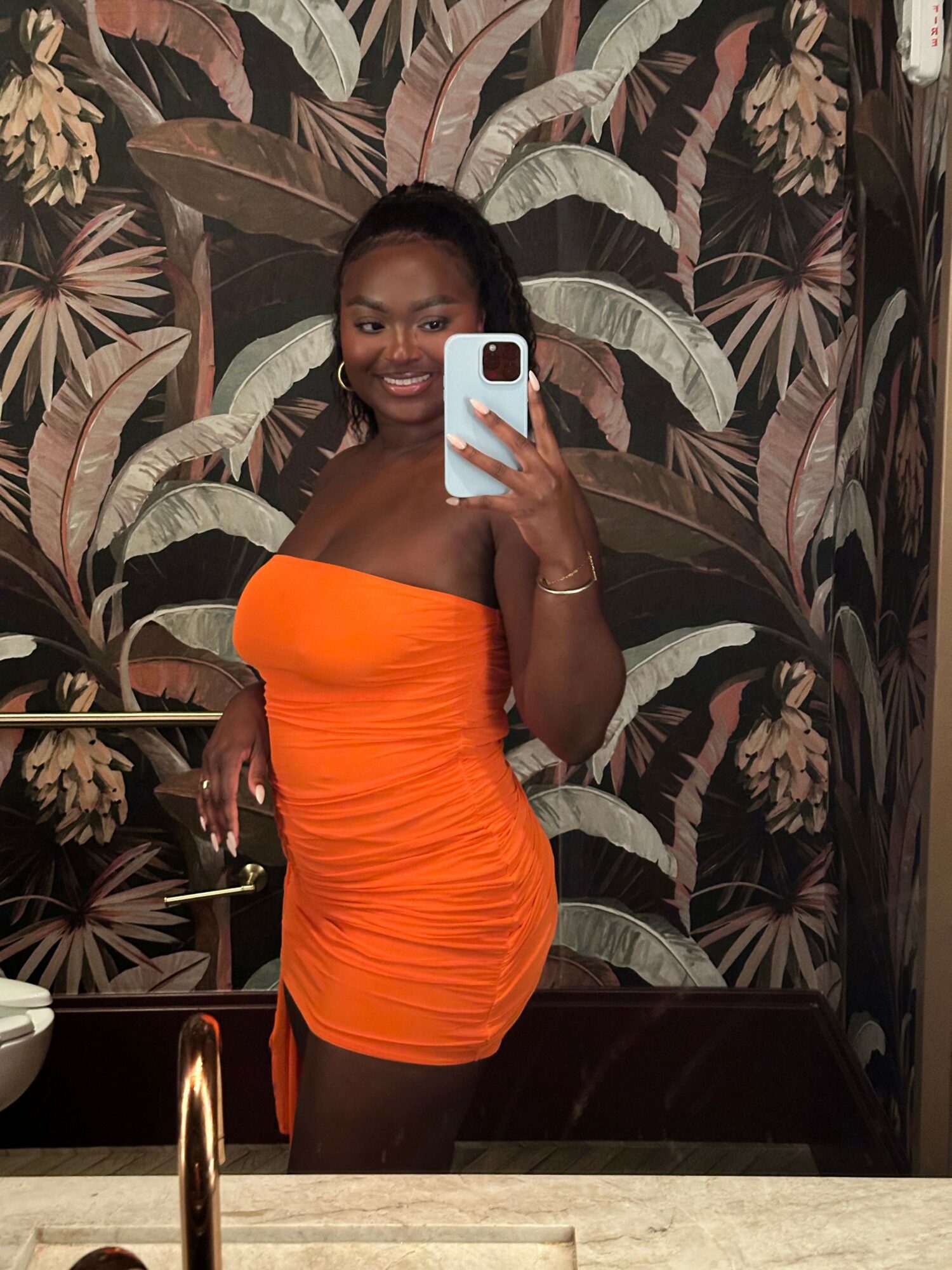 Woman in an orange dress taking a mirror selfie with a smartphone, standing in front of a floral-patterned wall.
