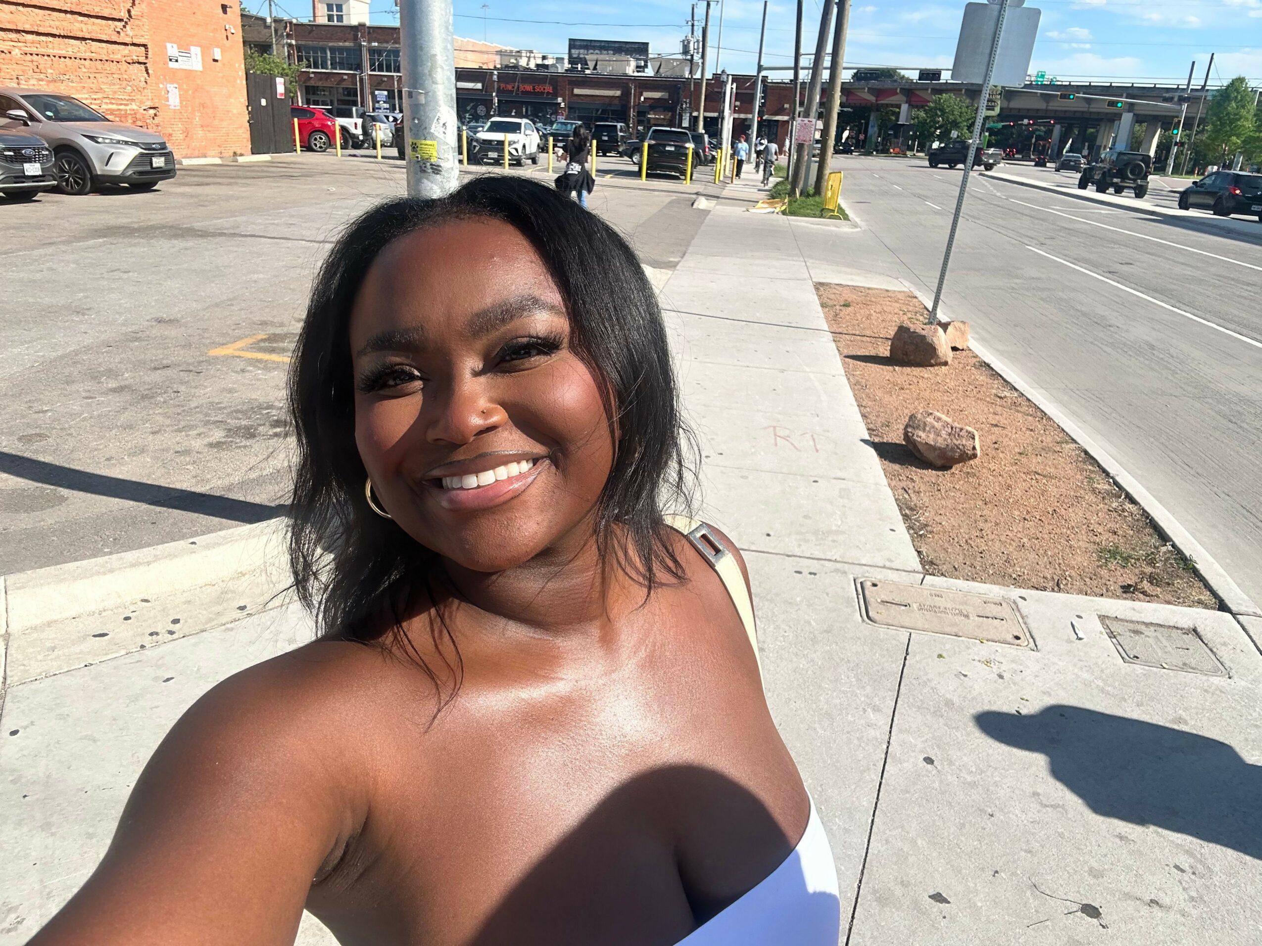 Smiling woman with dark hair outdoors, sunlight on her face, concrete sidewalk background.