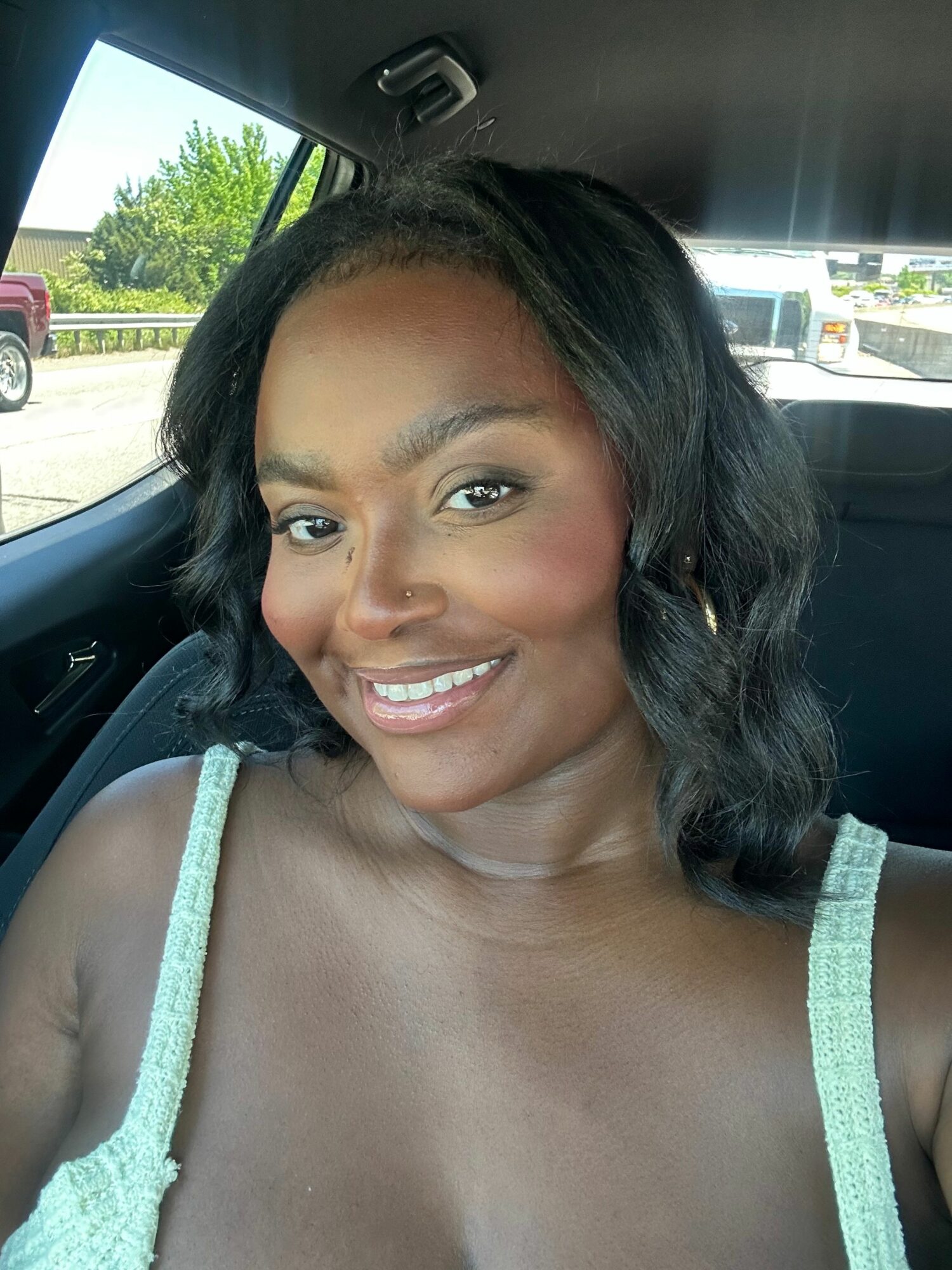 Young woman with dark hair smiling in a car, wearing a light-colored top, with trees and a building outside.