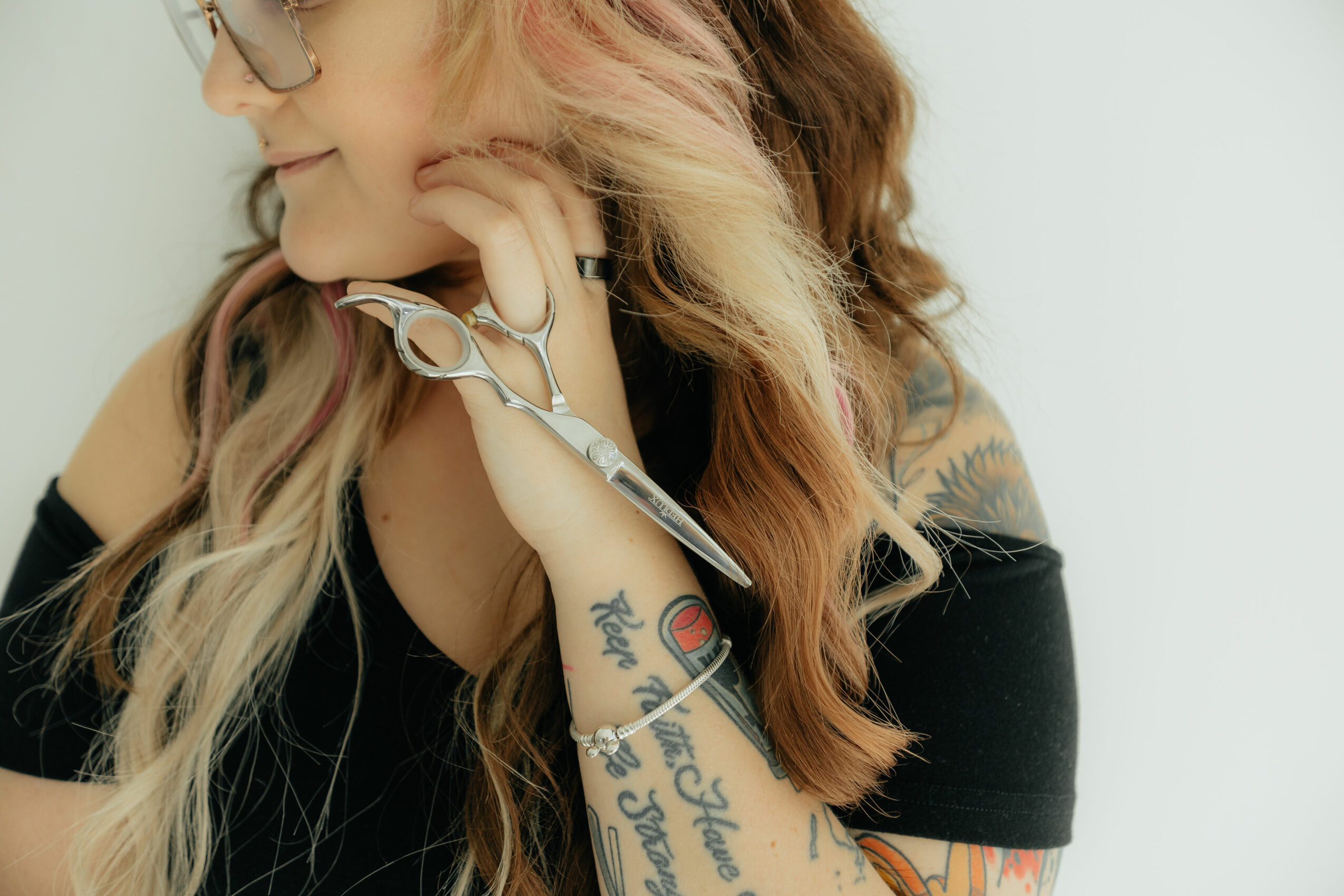 Woman with long, wavy hair holding scissors near her face, wearing glasses and a black off-shoulder top.