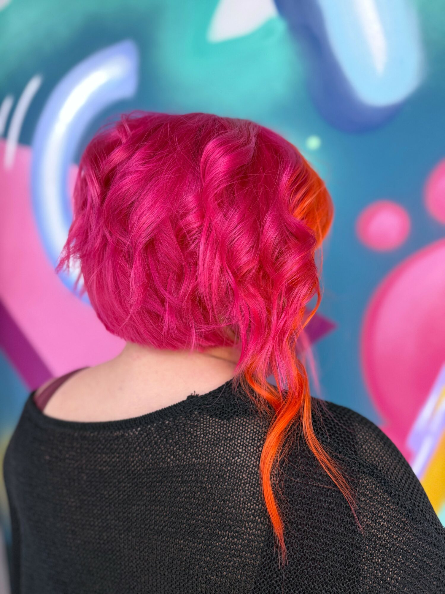 Person with vibrant pink and orange wavy hair facing away from camera, standing in front of colorful abstract background.