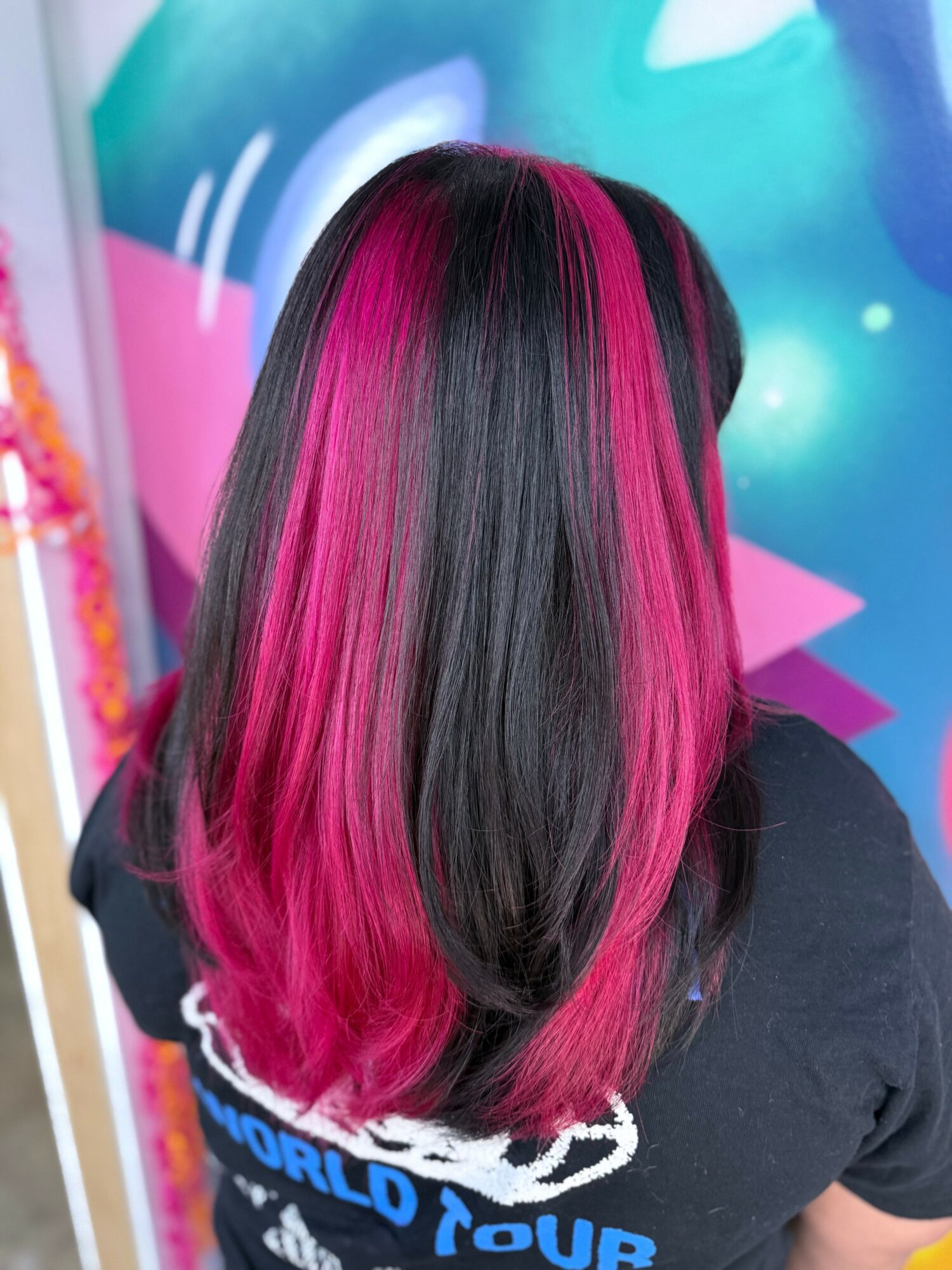 Person with long black hair and vibrant pink highlights, standing in front of a colorful mural.
