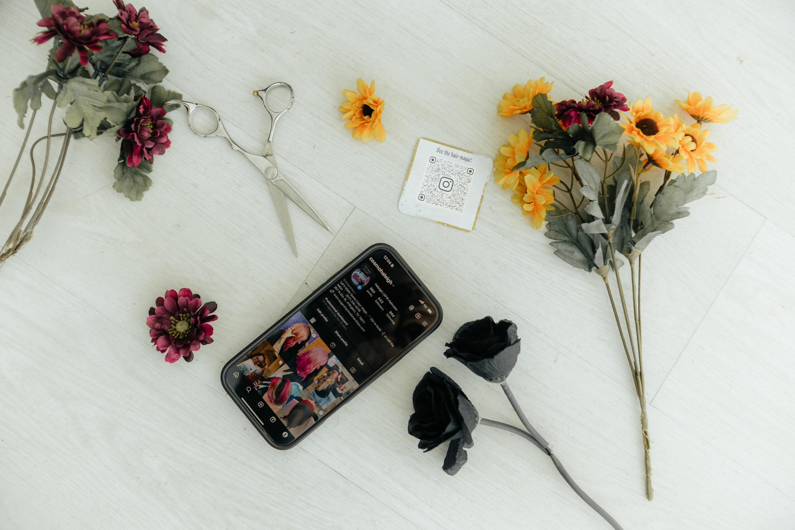 Flowers, a smartphone displaying a social media profile, a pair of scissors, and a small card on a white surface.