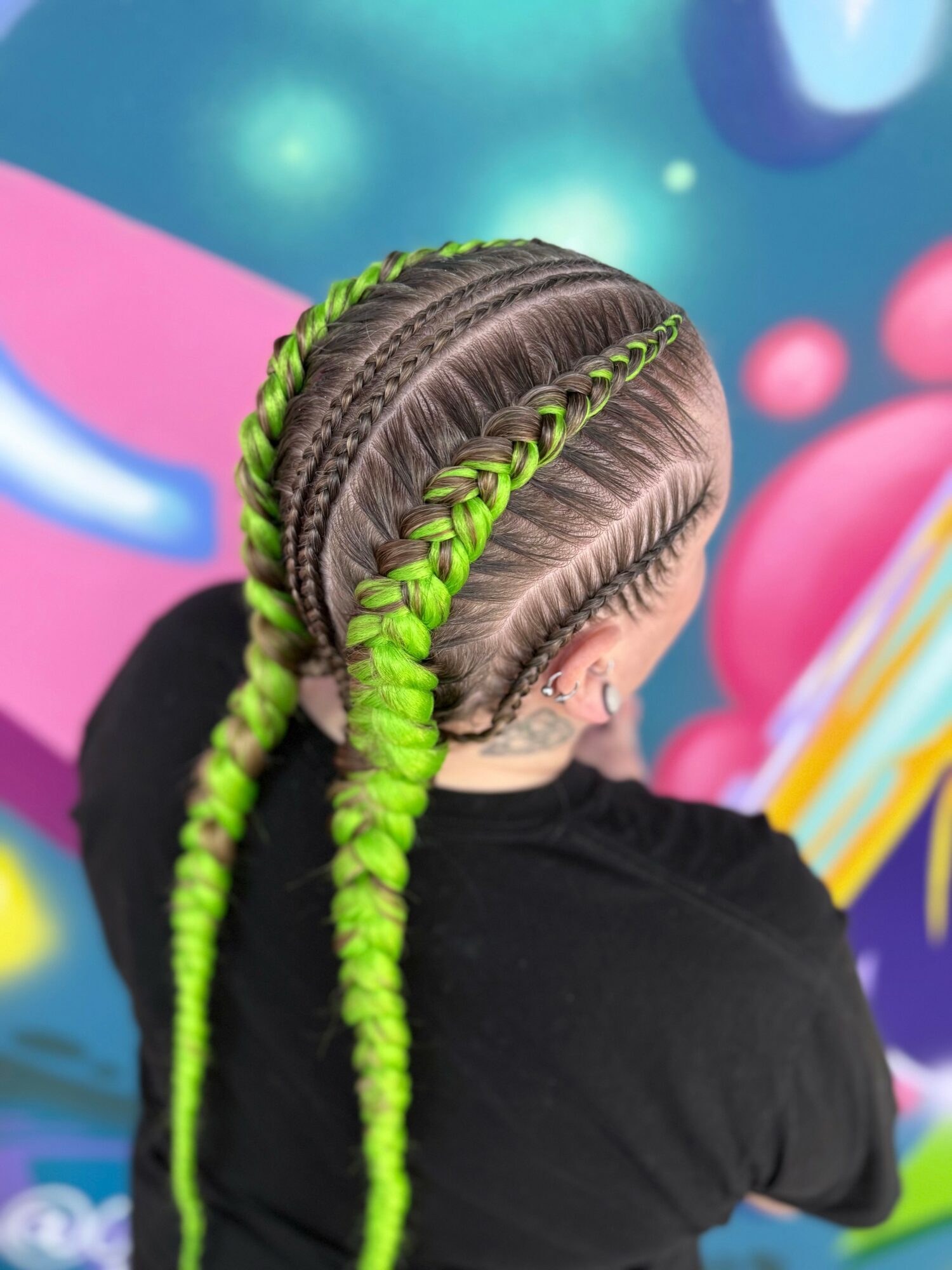 Person with braided hairstyle featuring green and black braids, facing away, against colorful background.