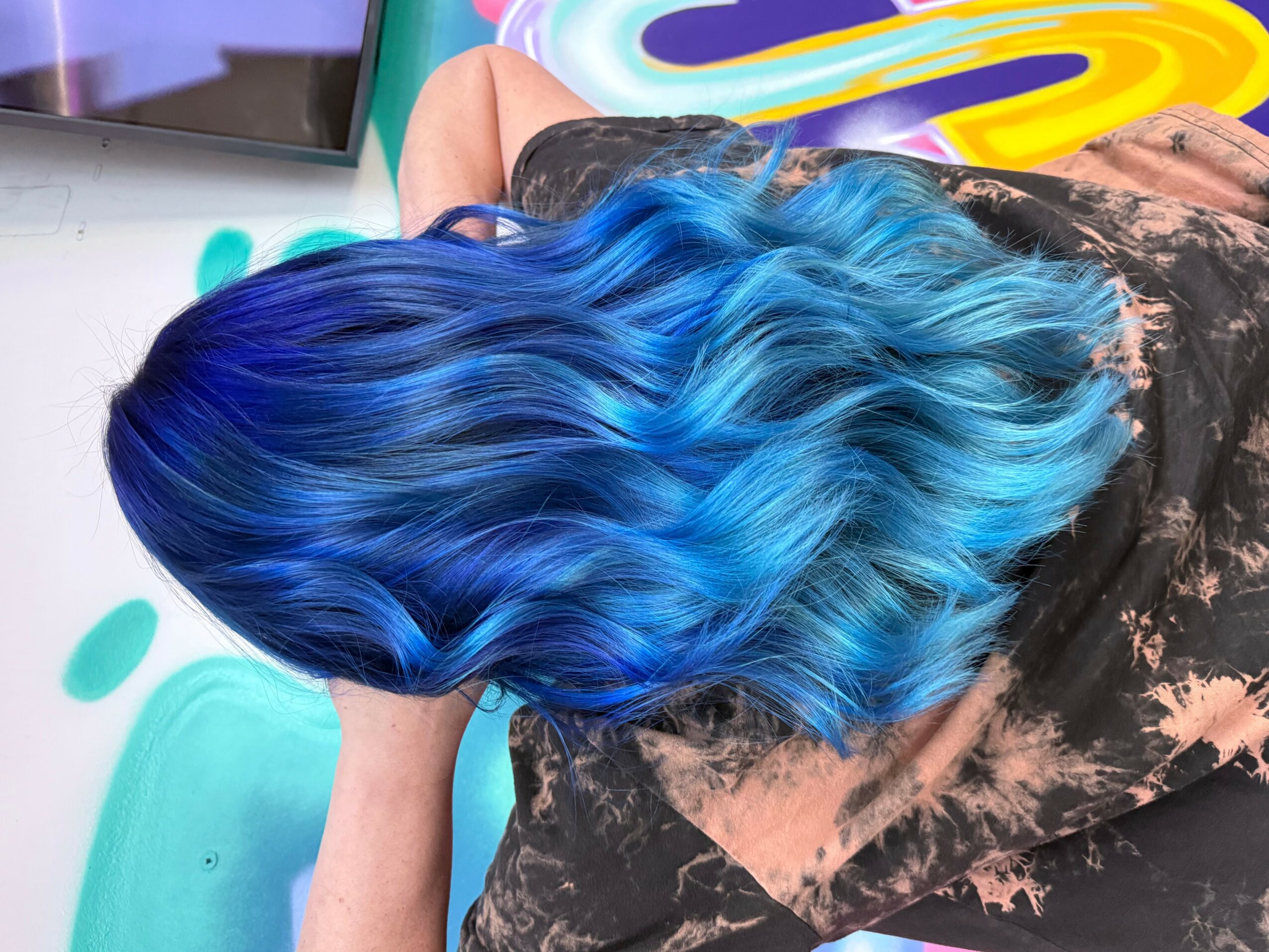 Person with long, wavy hair dyed in shades of blue, sitting with head down, wearing a dark patterned shirt.