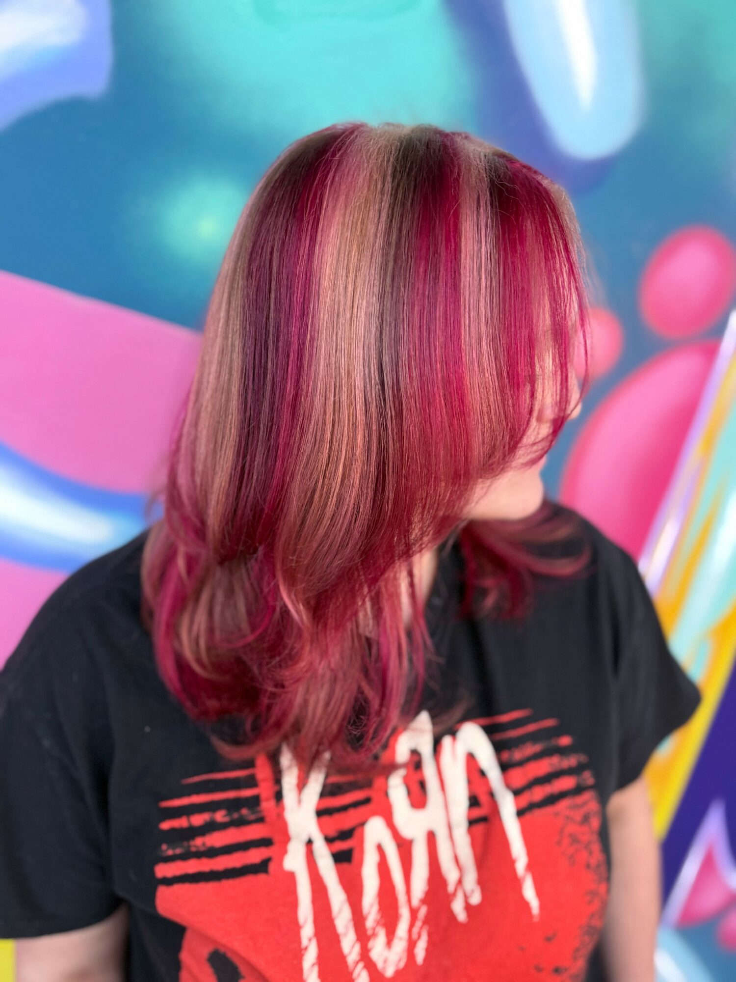 Person with multi-colored pink and blonde hair, wearing a black shirt with red and white text, against a colorful background.