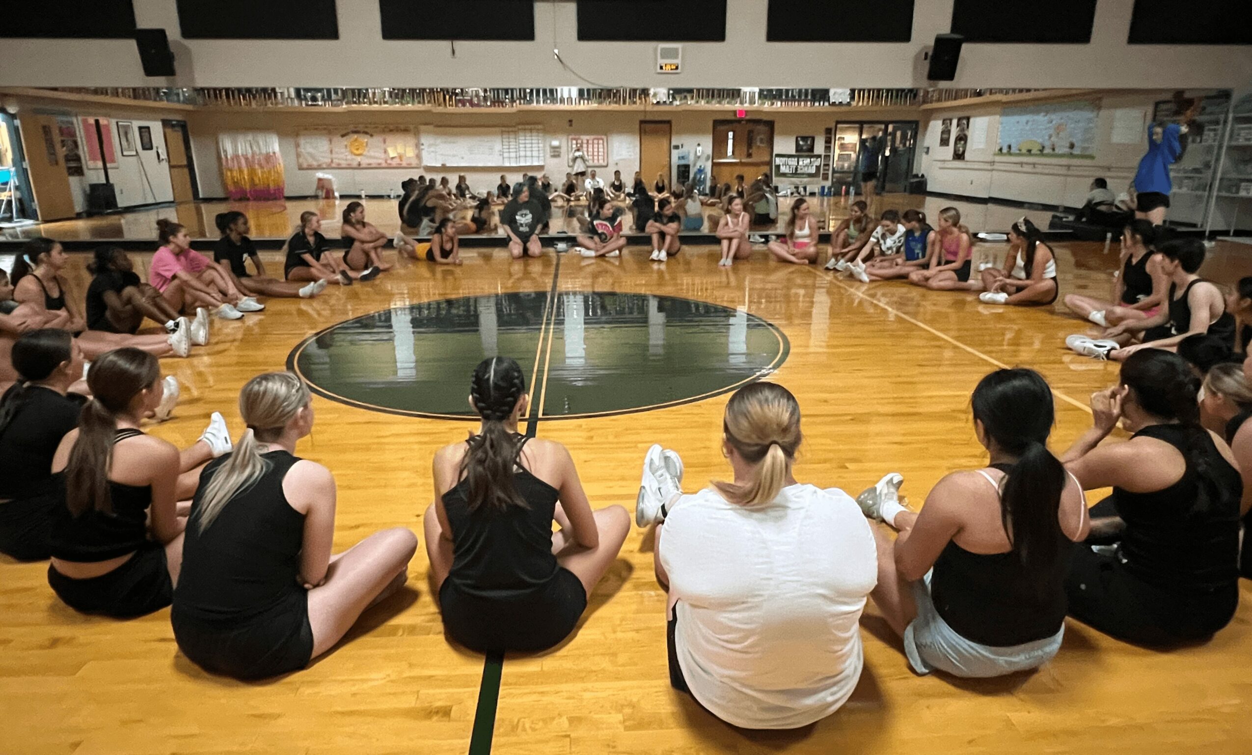 Group of people sitting in a circle on a wooden floor in a gym or dance studio, with a mirror wall and a fountain in the center.
