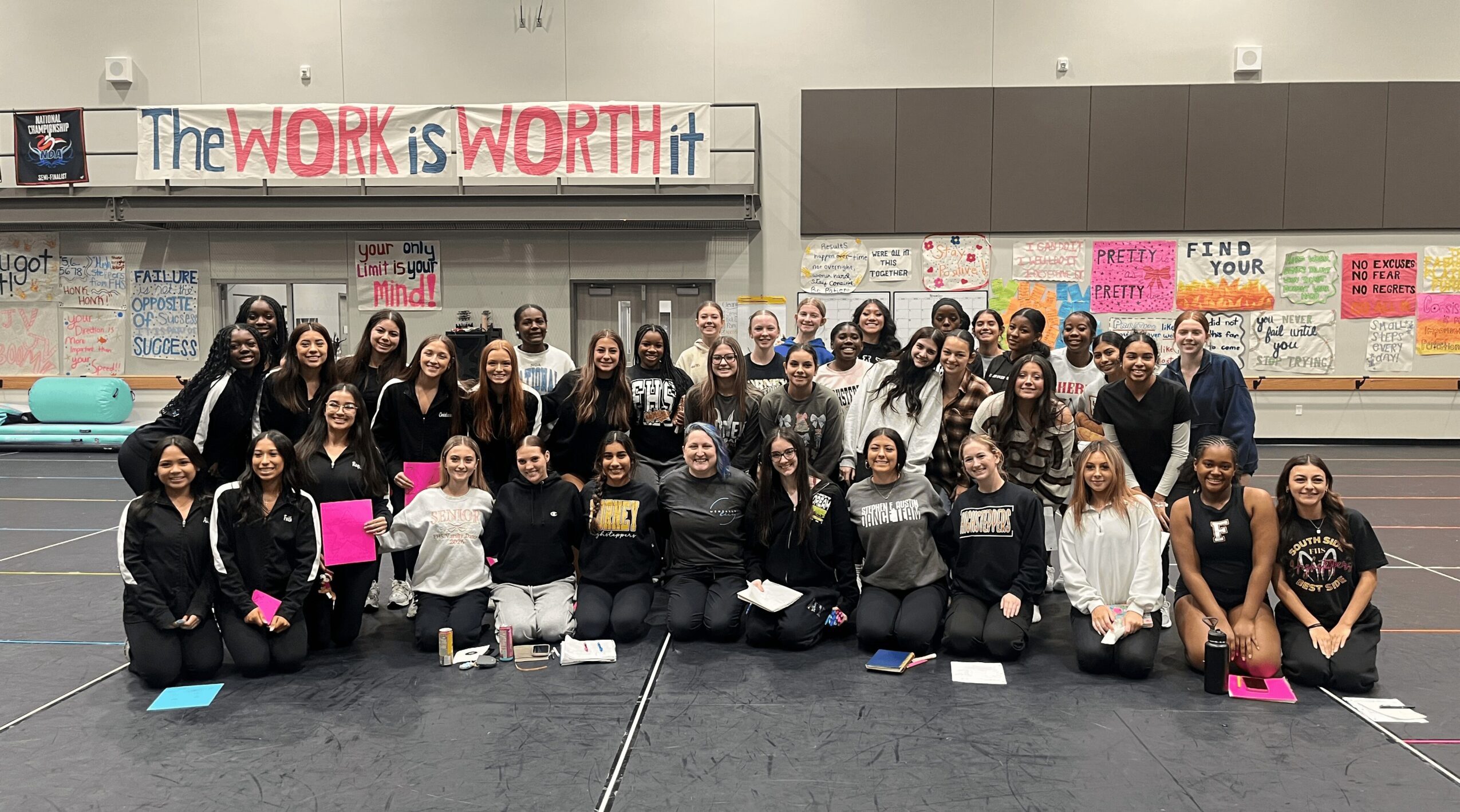 Group of people in a gymnasium holding signs, with a banner reading 'The WORK is WORTH it' behind them.