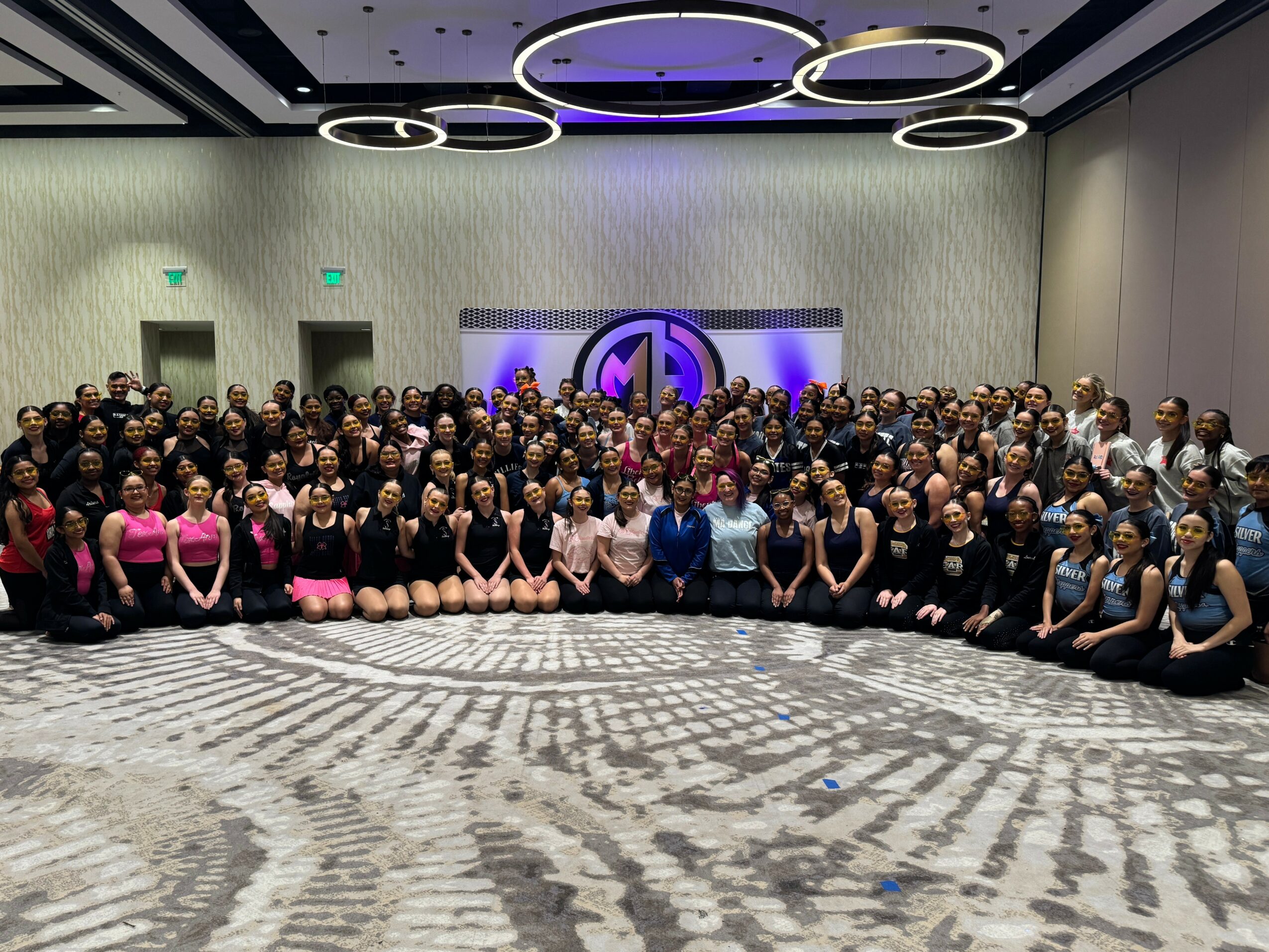 Large group of people in a conference room, some wearing pink, posing for a photo with a logo on the wall behind.