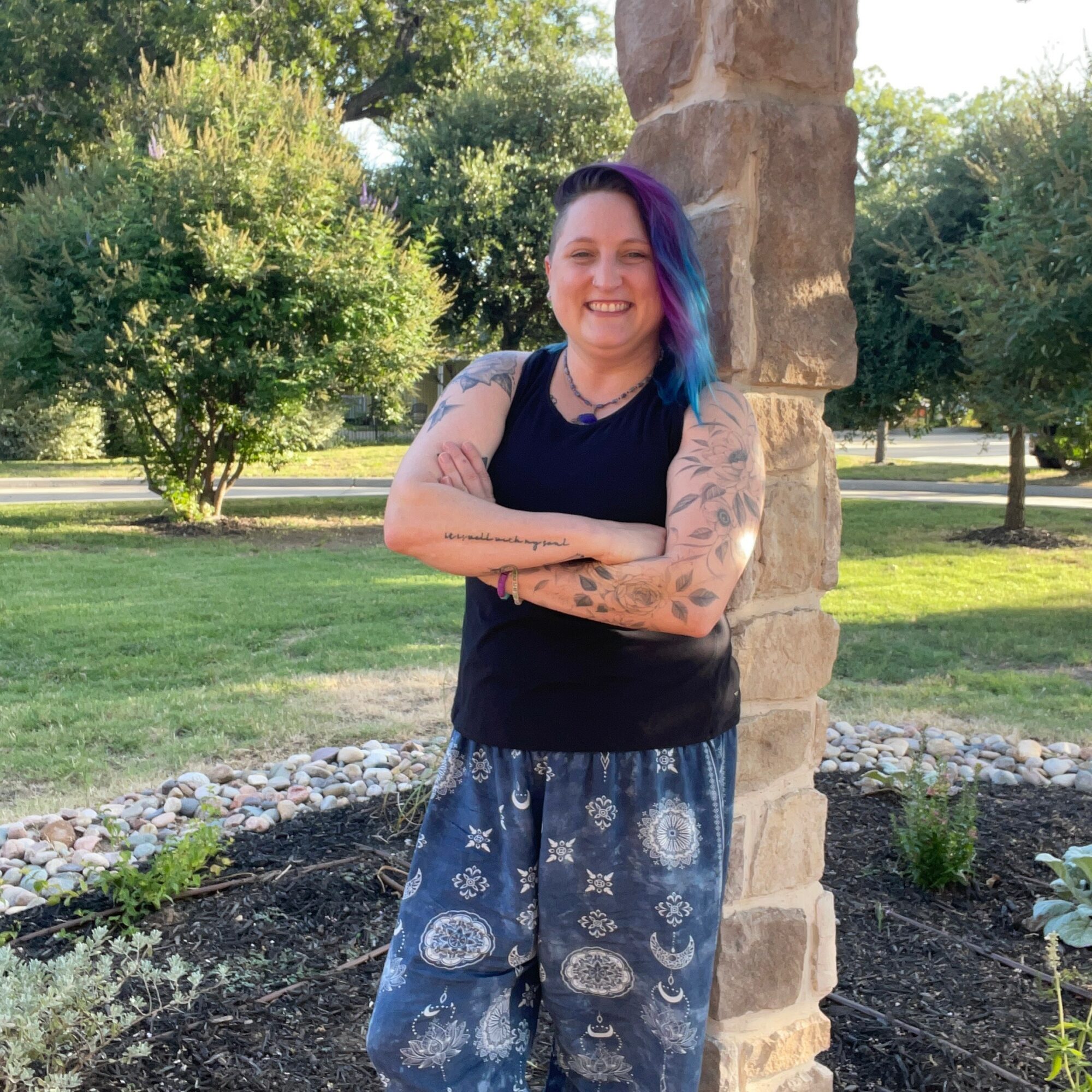 Smiling person with colorful hair and tattoos, standing outdoors next to a stone pillar, arms crossed, in a park.