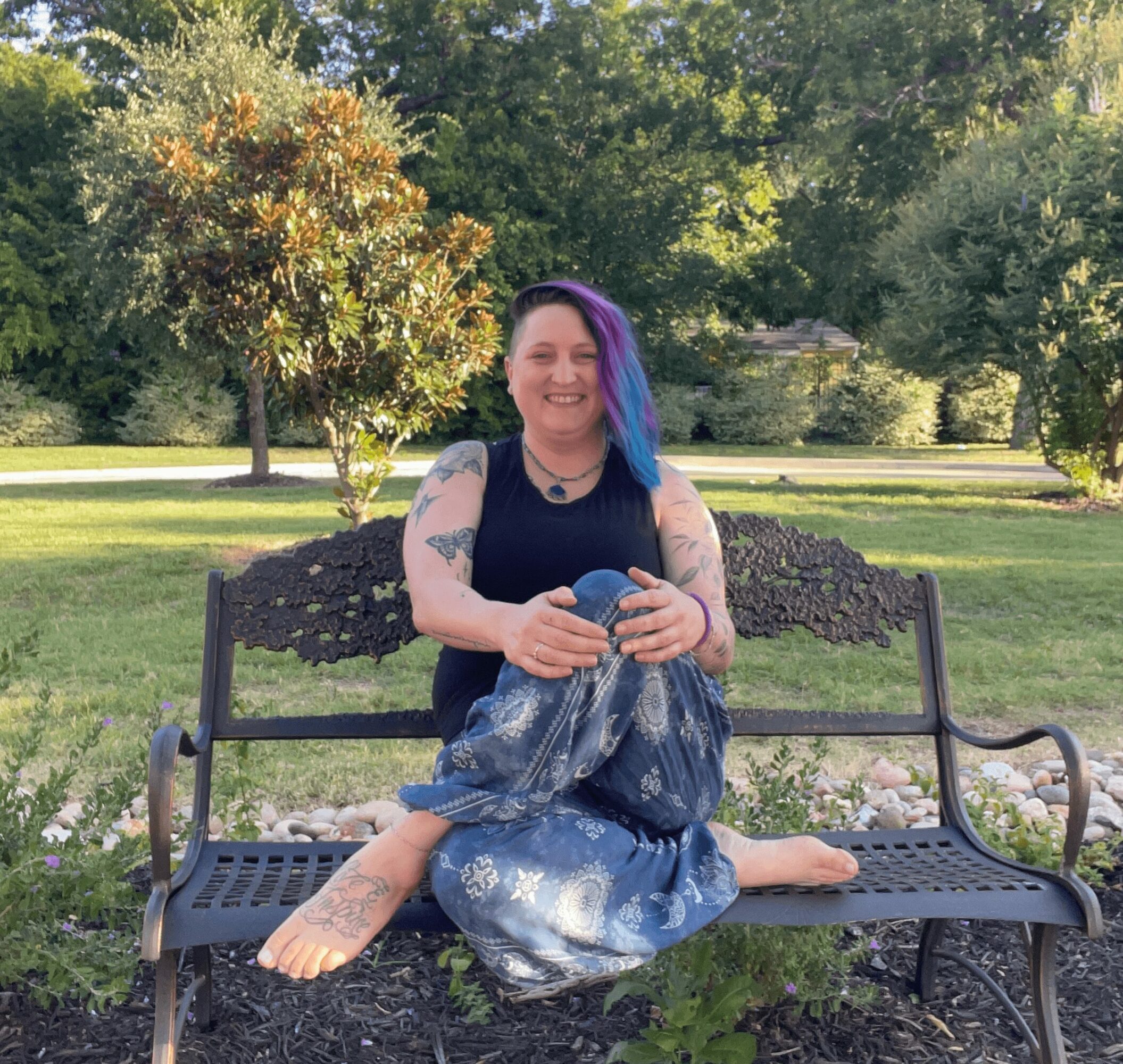Person with colorful hair sitting cross-legged on a park bench, smiling, surrounded by trees and greenery.