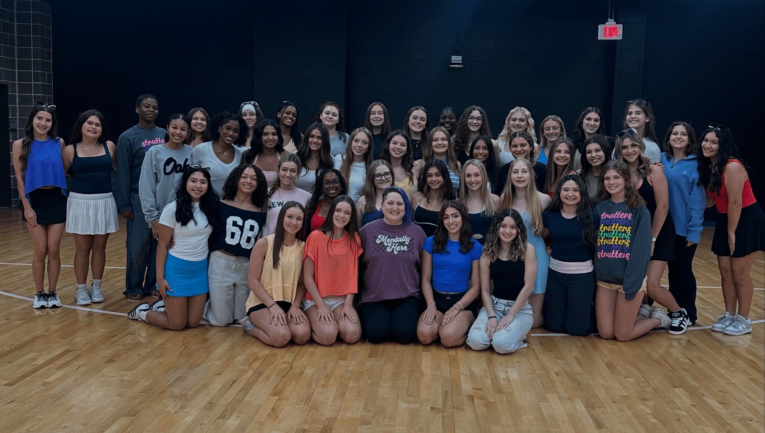 Group of young women and girls standing in a dance studio with a wooden floor and black walls.