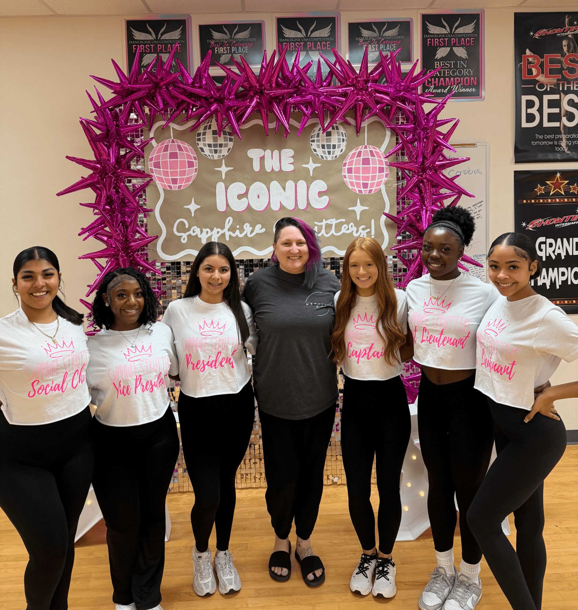 Group of seven women standing in front of a pink heart-shaped decoration with text, smiling, indoors.