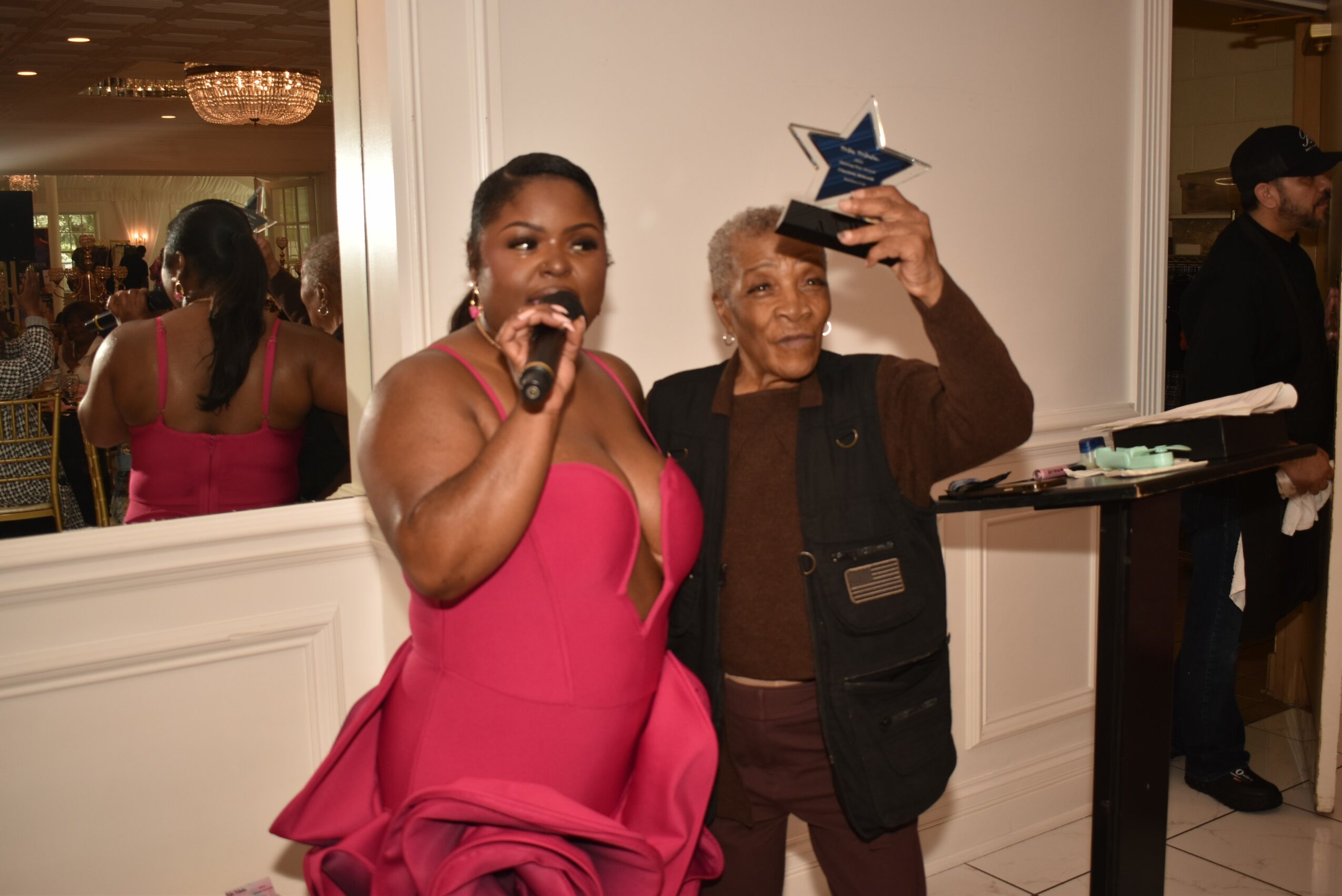 Two women at an event, one holding a microphone and the other holding a star-shaped award, smiling and celebrating.