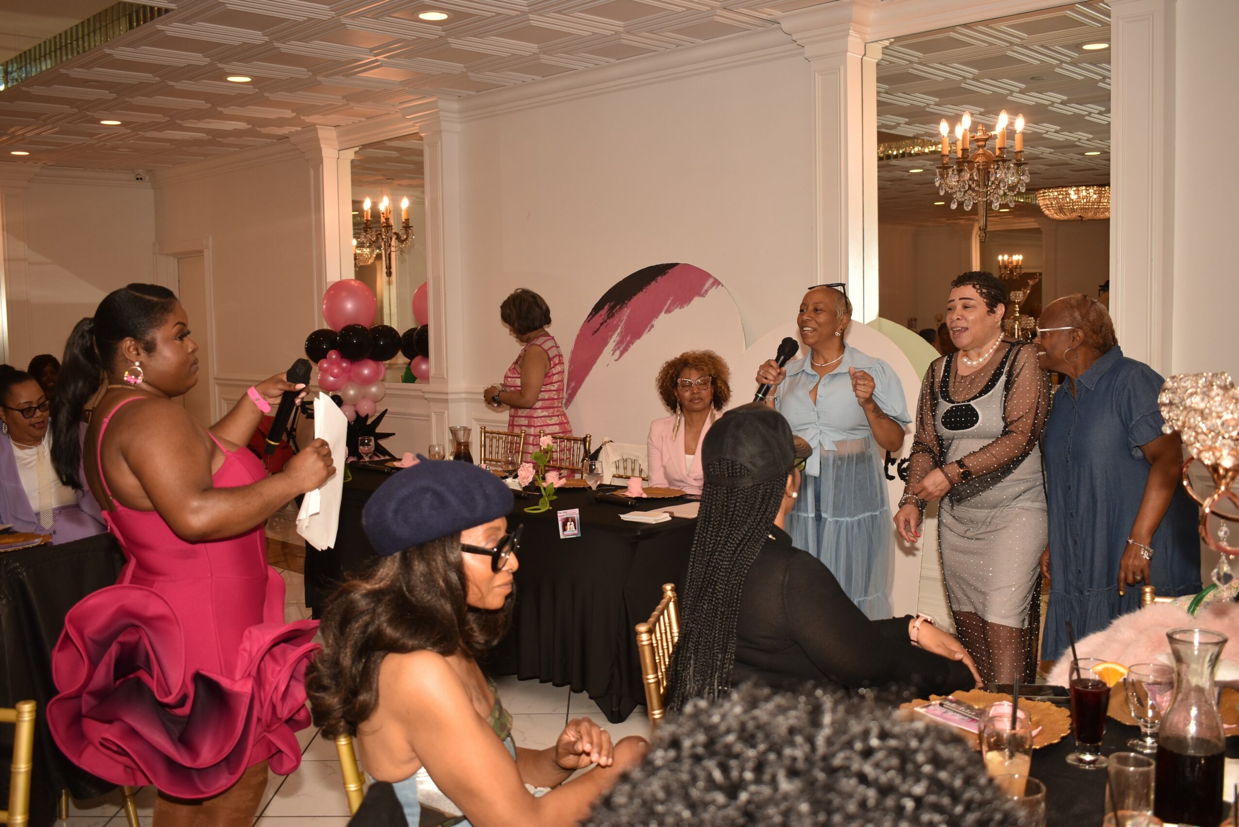 Group of women gathered in a decorated room, some standing and some sitting, with tables and balloons visible.