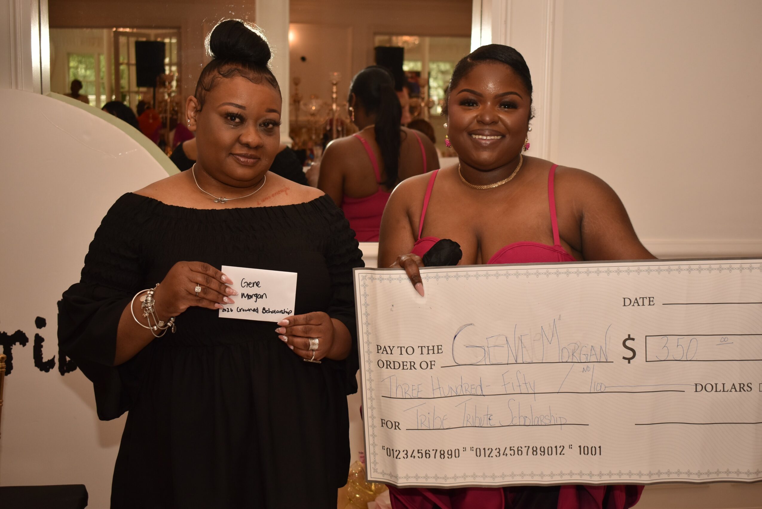 Two women smiling, one holding a check and the other holding a large check, in an indoor setting.