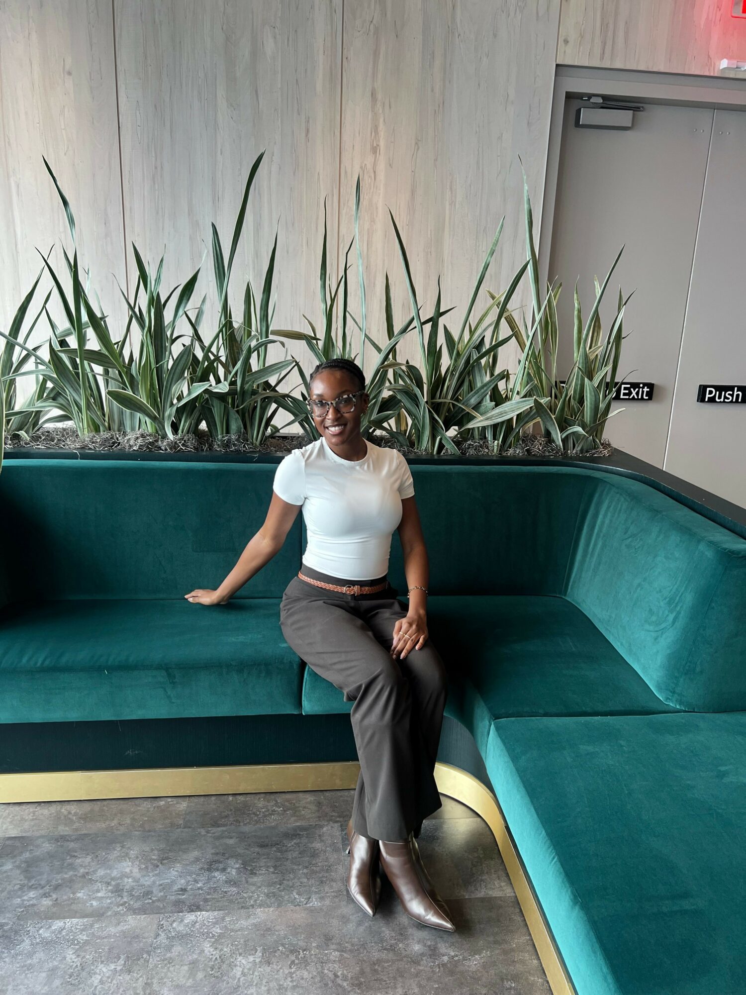 Woman in white top and gray pants standing next to a large green planter with tall plants, indoors.