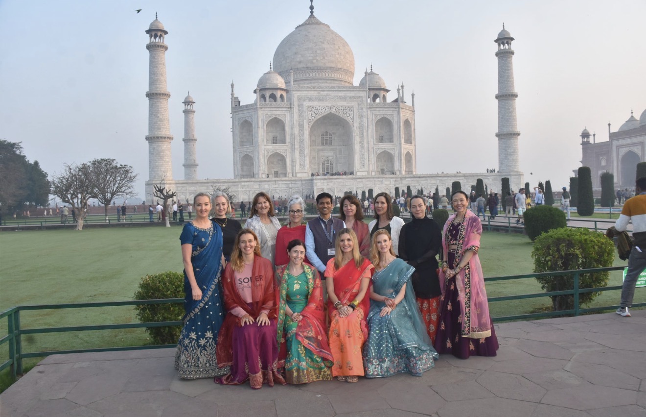 Group of people in colorful traditional clothing standing in front of the Taj Mahal, with green lawns and trees behind.
