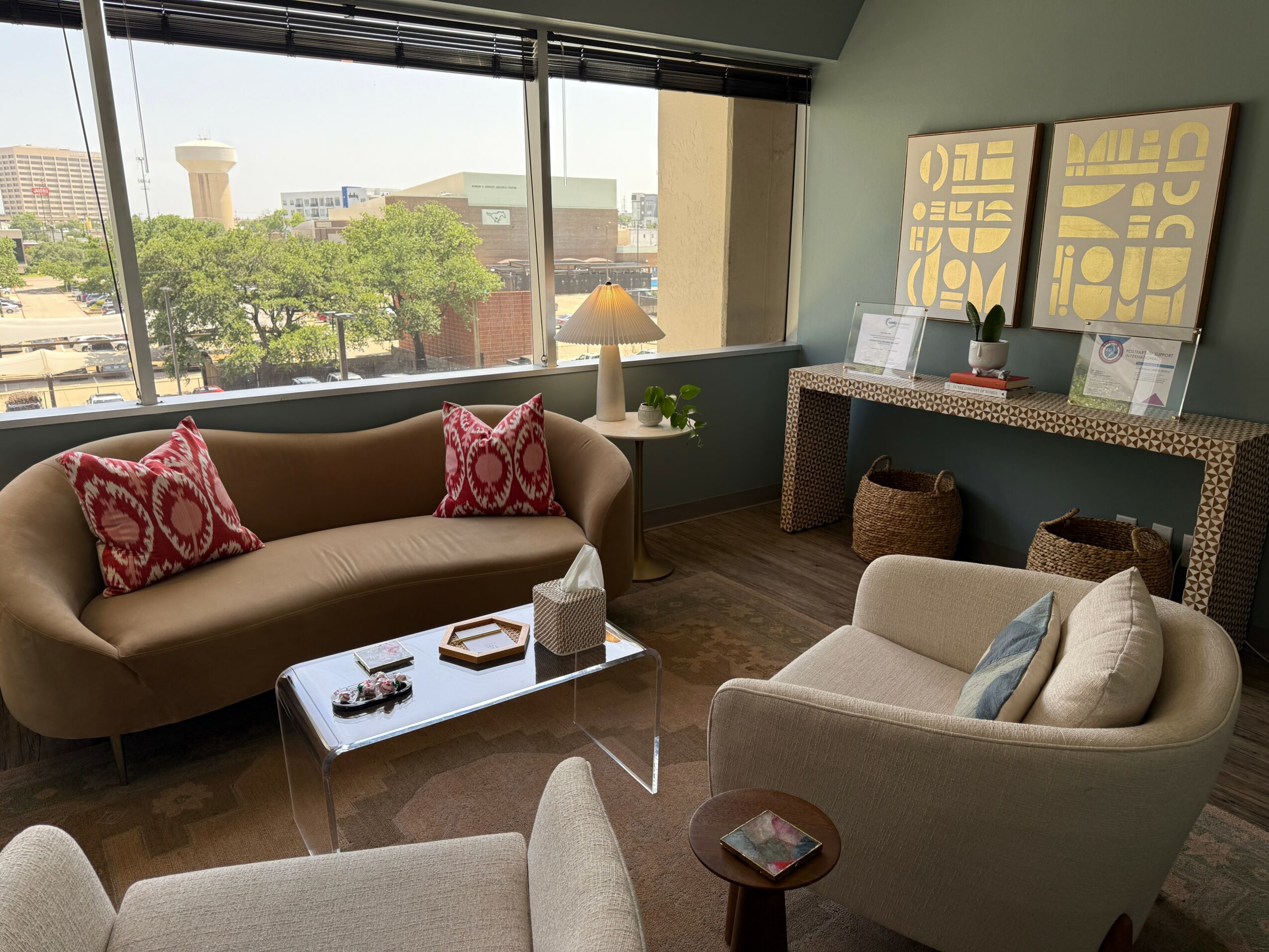 Living room with beige sofa, armchair, coffee table, and large window showing cityscape, trees, and buildings.