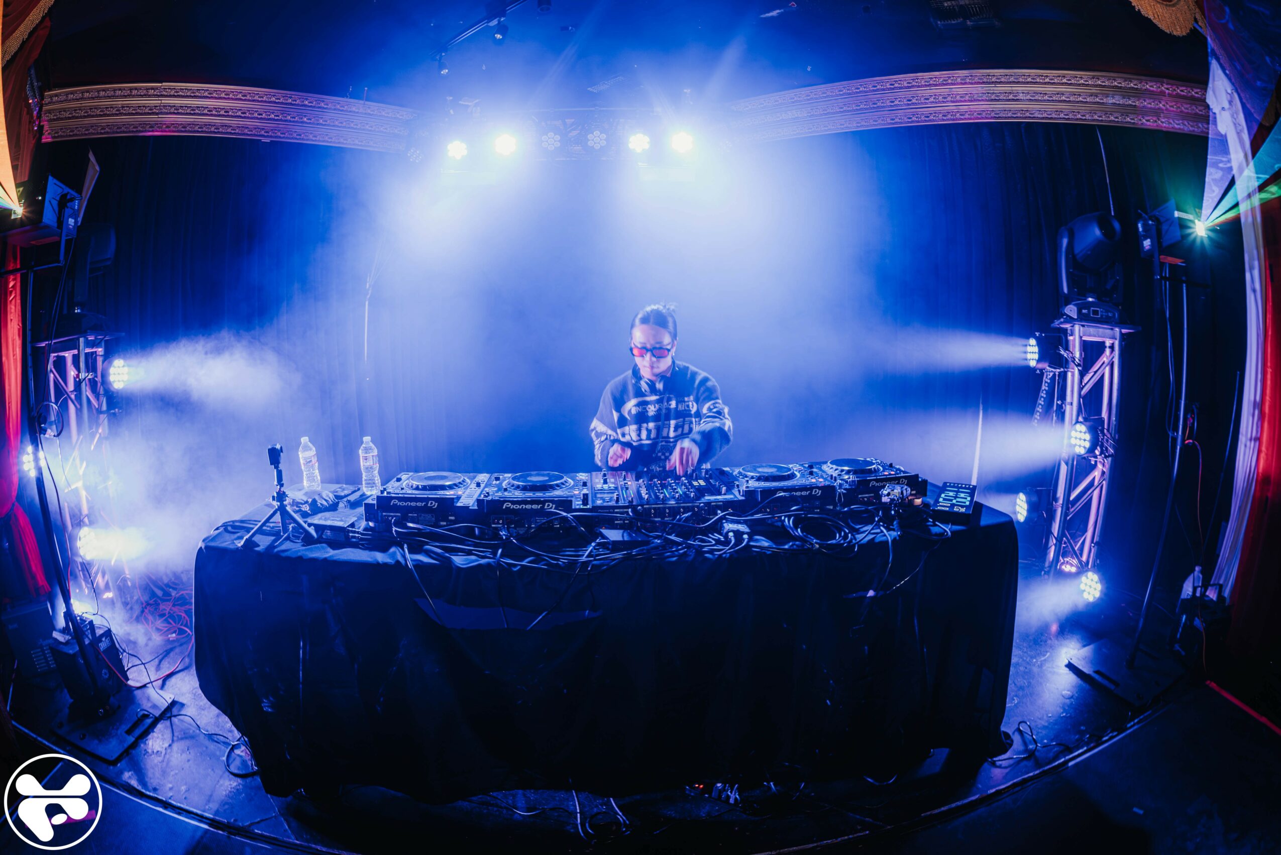 DJ stands behind a mixing console on stage with blue and purple lighting, fog, and equipment, in a concert setting.