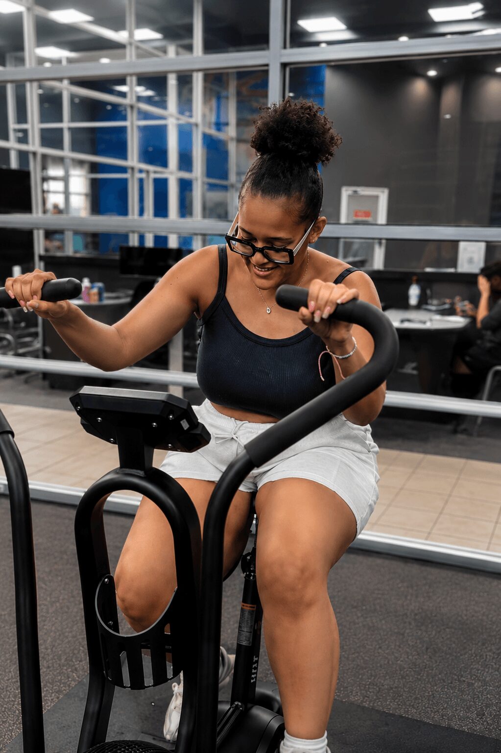 Woman exercising on stationary bike in gym, wearing glasses, black tank top, and white shorts, with large windows behind her.