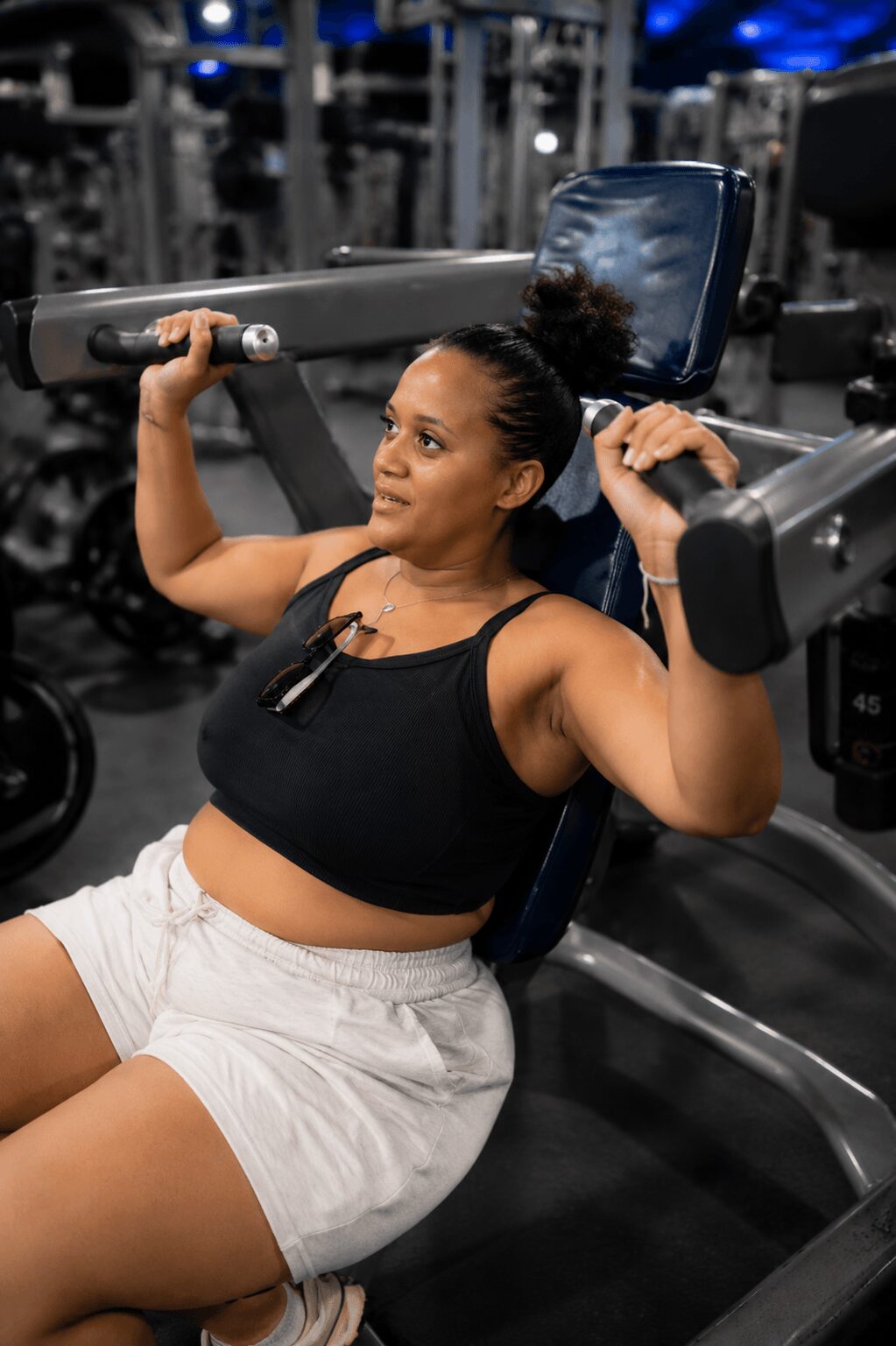 Woman lifting a barbell in a gym, seated on a workout machine, wearing a black sports top and white shorts.