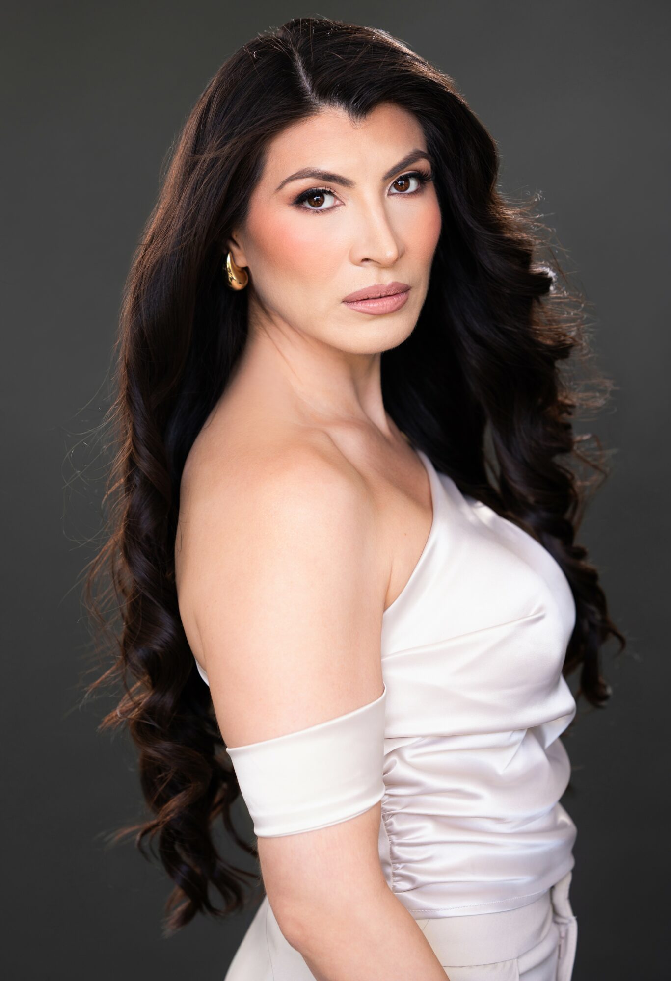 Woman with long dark hair wearing a white off-shoulder top, posing against a dark background.