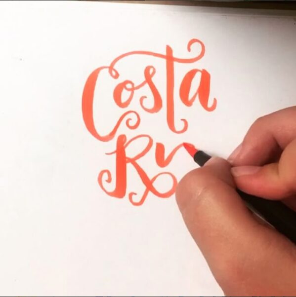 Hand writing the word 'Ostak' in orange cursive on white paper.