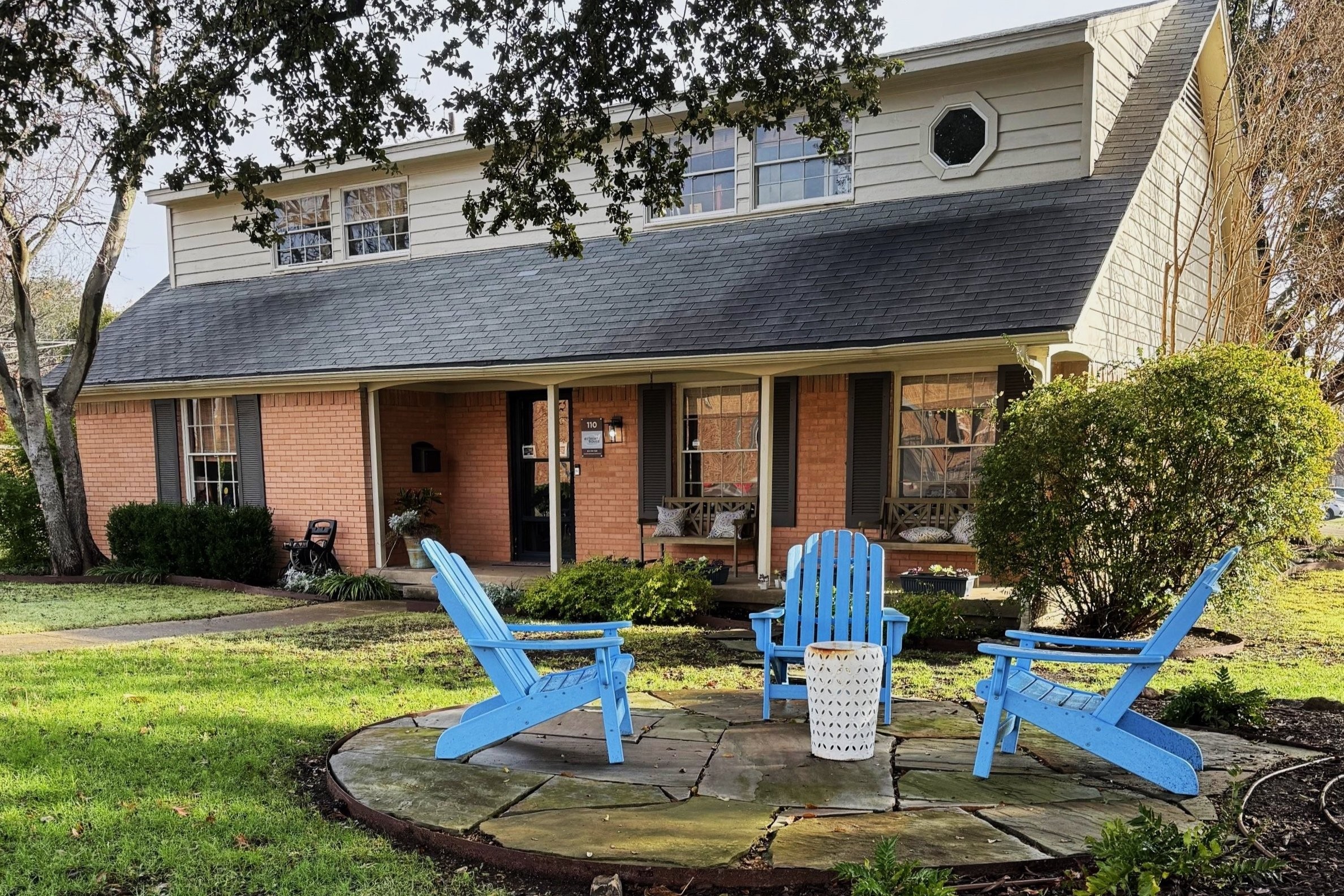 Front yard with two blue Adirondack chairs, a small table, and a house with brick and siding exterior, surrounded by trees and bushes.