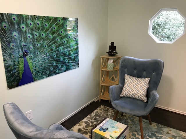 Living room corner with peacock artwork, gray armchair with pillow, small wooden shelf, octagonal window, and patterned rug.