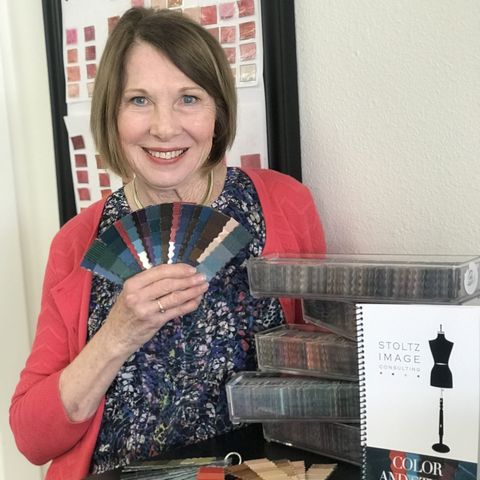 Woman with short brown hair smiling, holding color swatches, standing next to stacked boxes and a mannequin sketch, in a room.