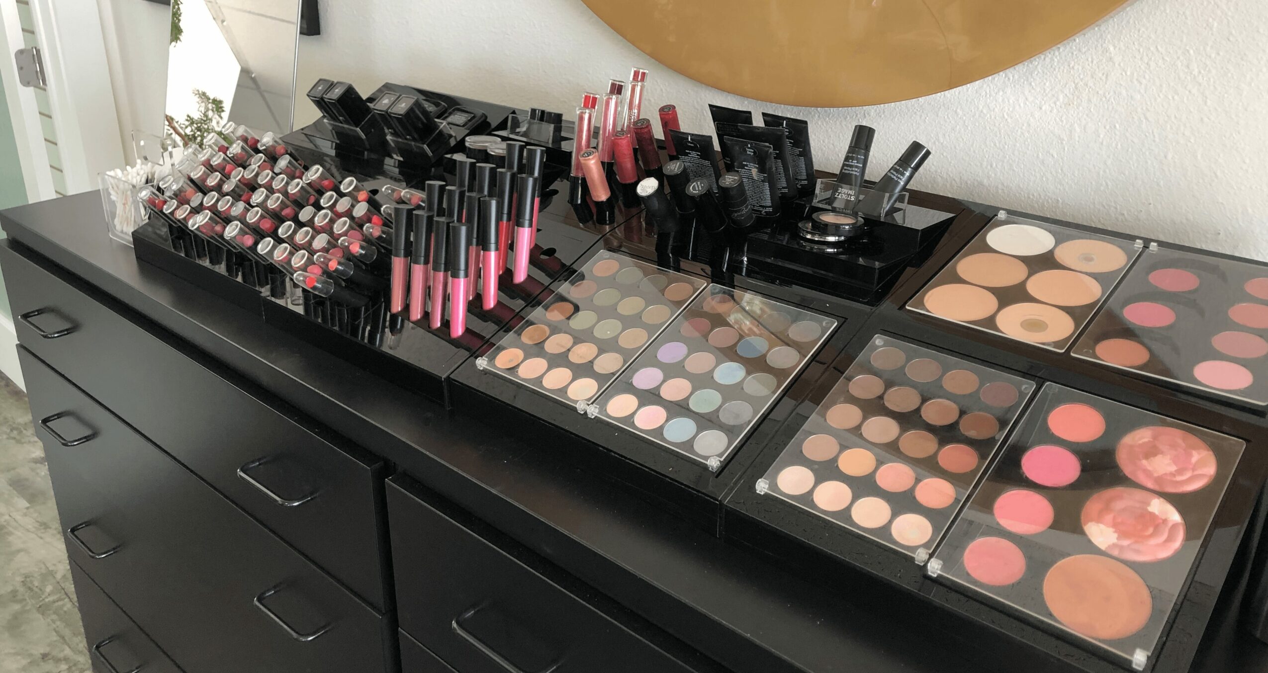 Makeup products including lipsticks, eyeshadows, and blushes arranged on a black surface, with drawers below and a wall behind.