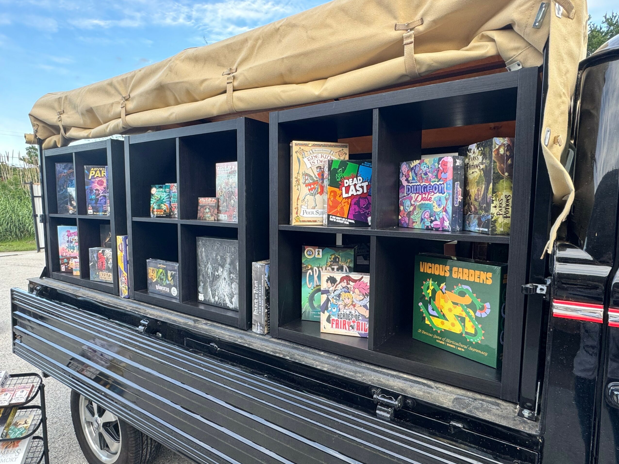 Black display shelf with toys and games on a truck bed, beige canopy above, outdoors with trees and sky visible.