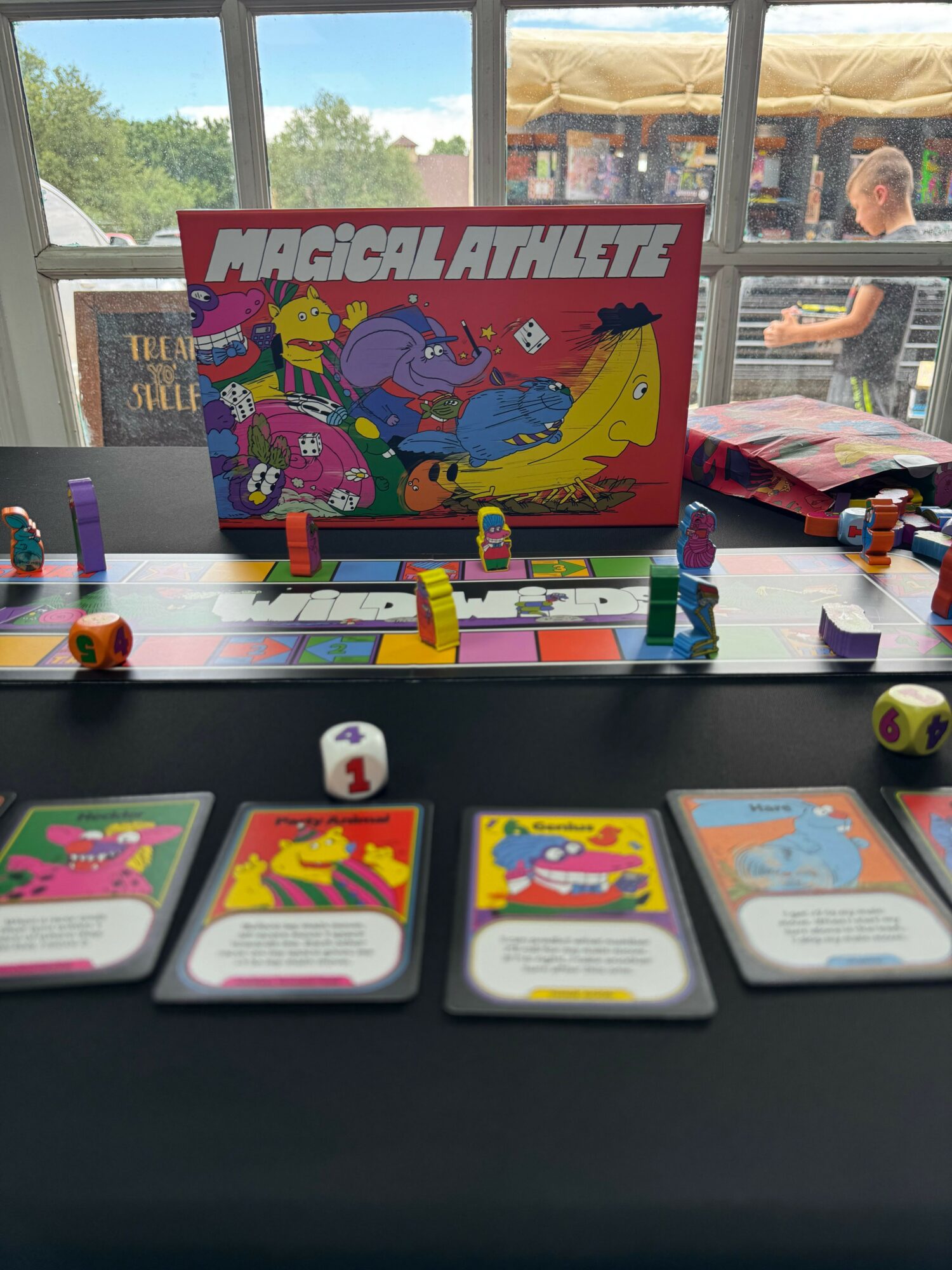 Board game with colorful cards and game pieces on a table, window with trees outside in background.