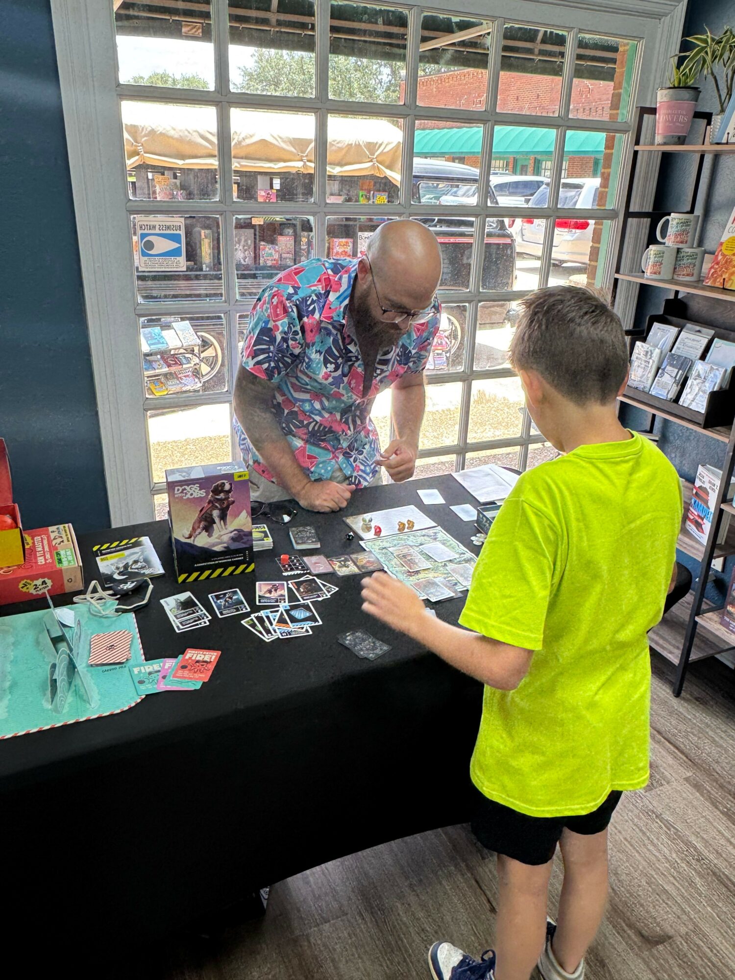 Two people browsing trading cards at a store, one standing and one sitting, with shelves and windows in the background.