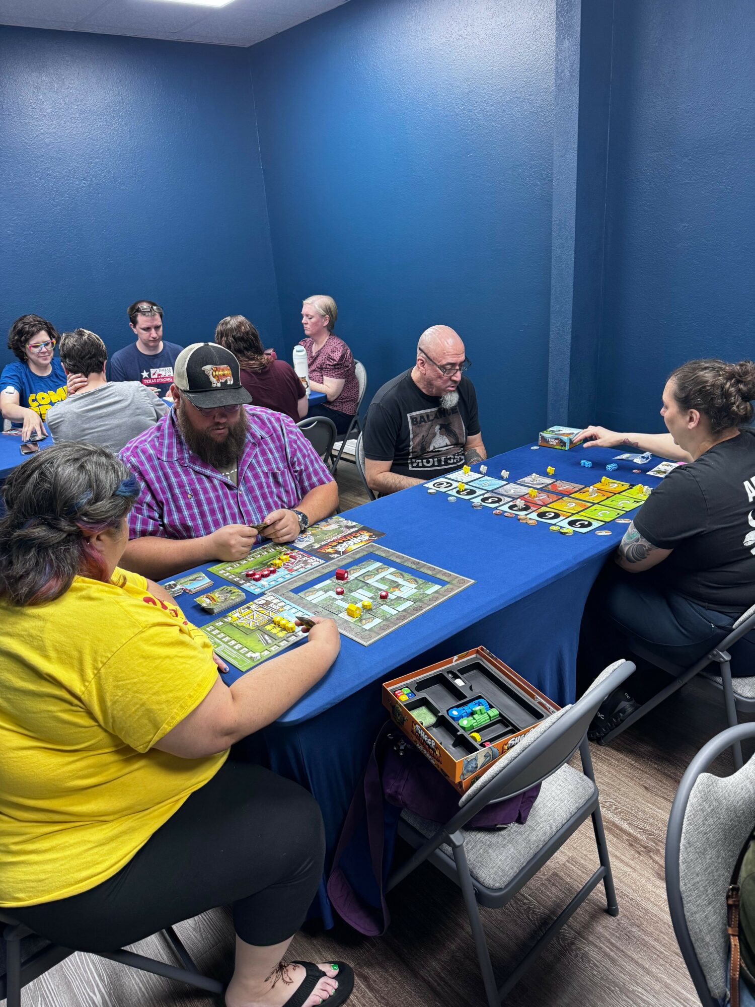 People playing a board game at a table in a room with blue walls, some wearing masks, with game boxes and cards visible.