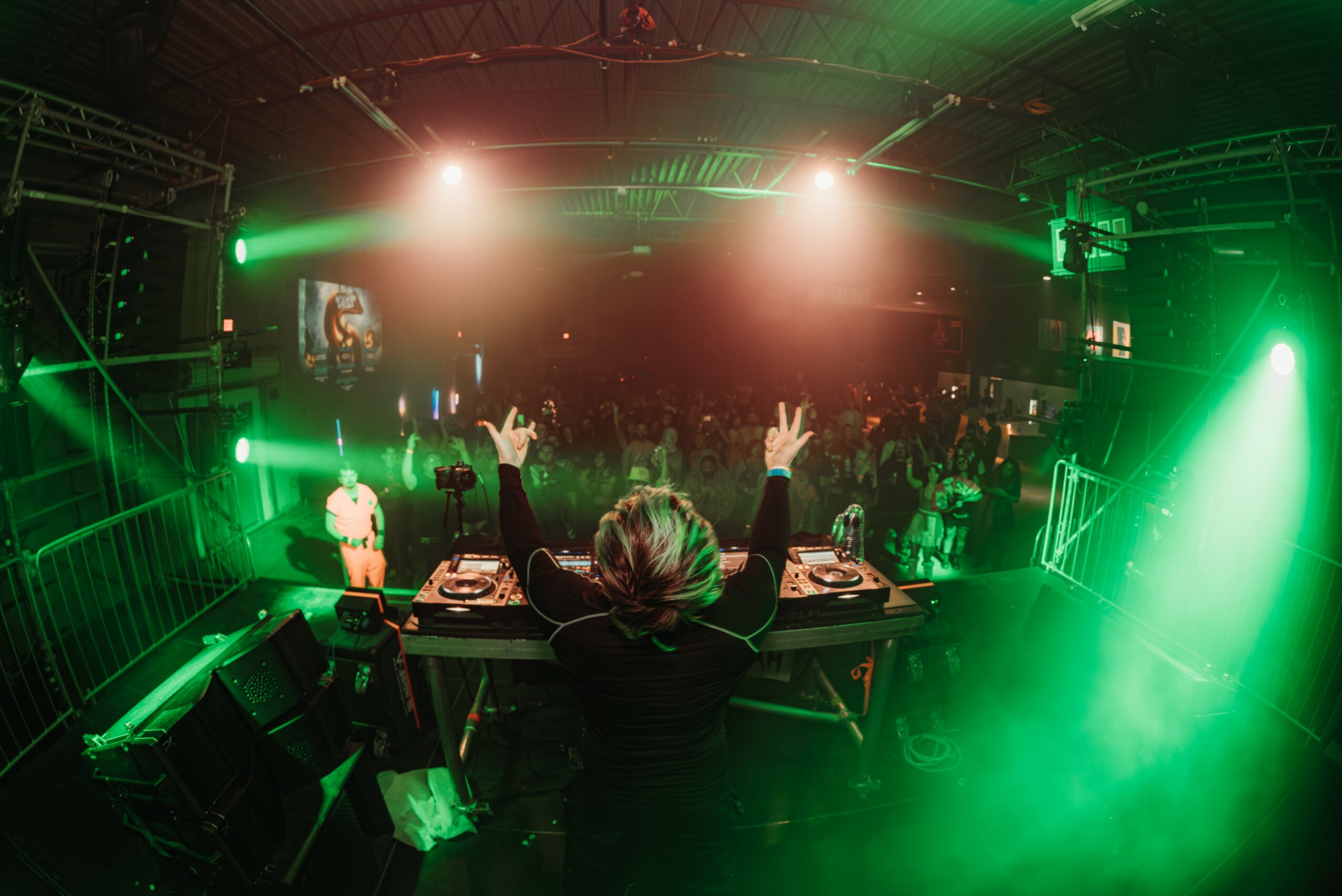 DJ with raised hands performing at a nightclub with green and pink lighting, crowd dancing in the background.
