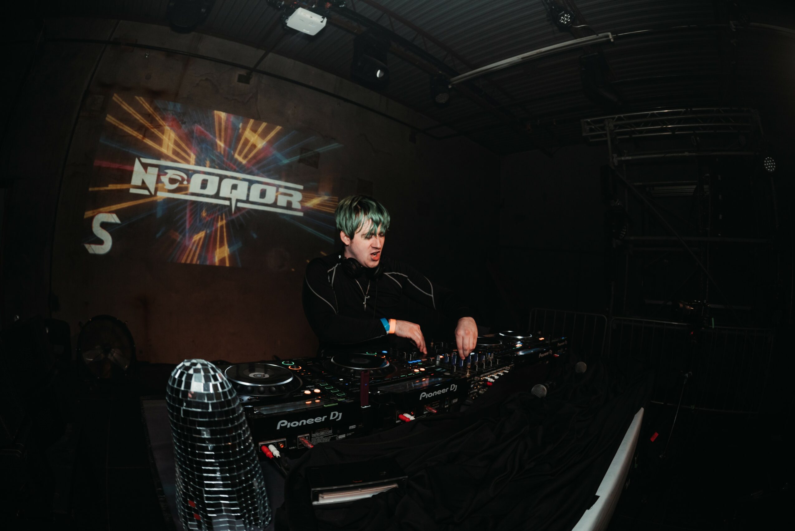 DJ with headphones operating turntables in a dark club with a logo projection on the wall.