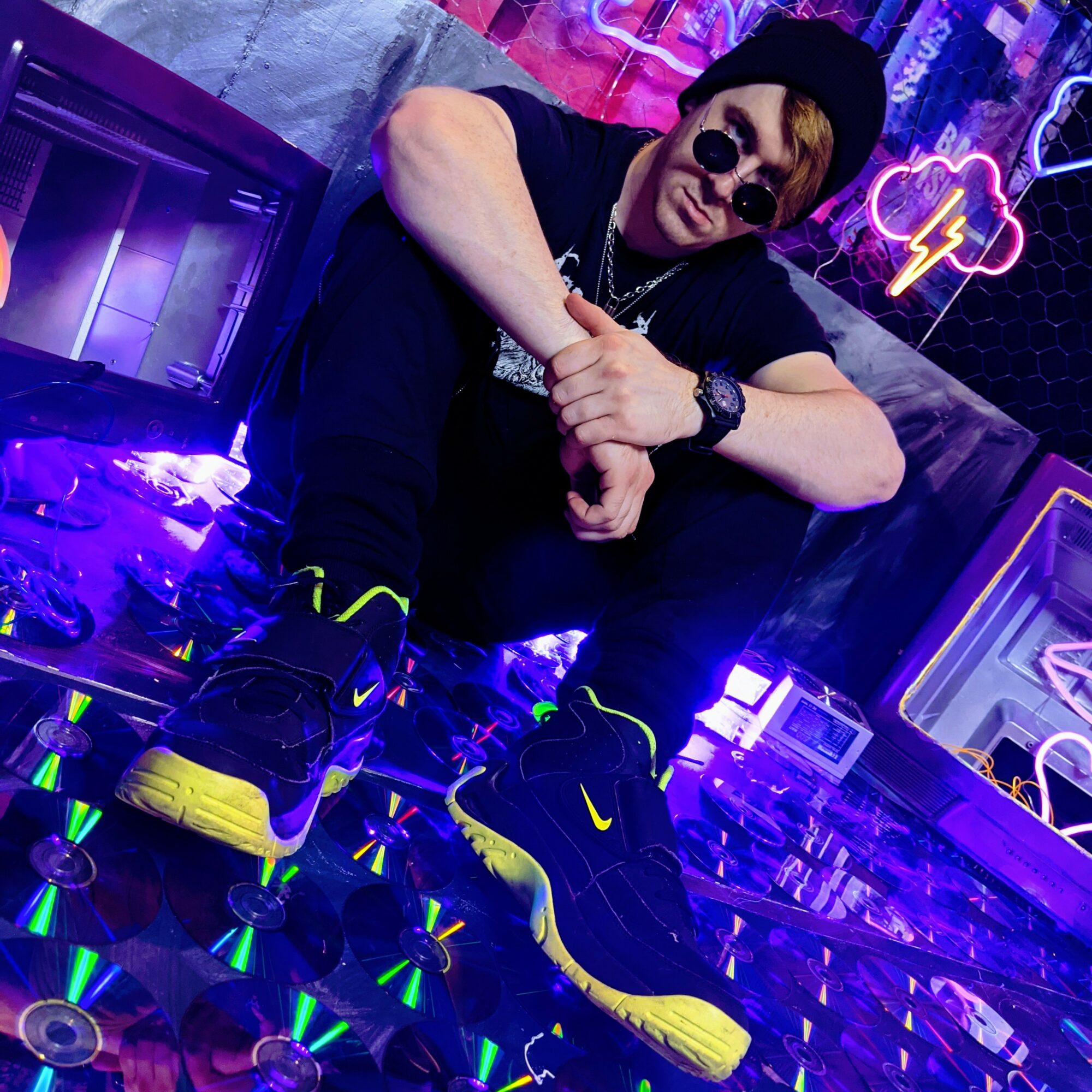Person sitting with crossed arms, wearing sunglasses, black clothing, and yellow-accented sneakers, in a neon-lit room with colorful signs.
