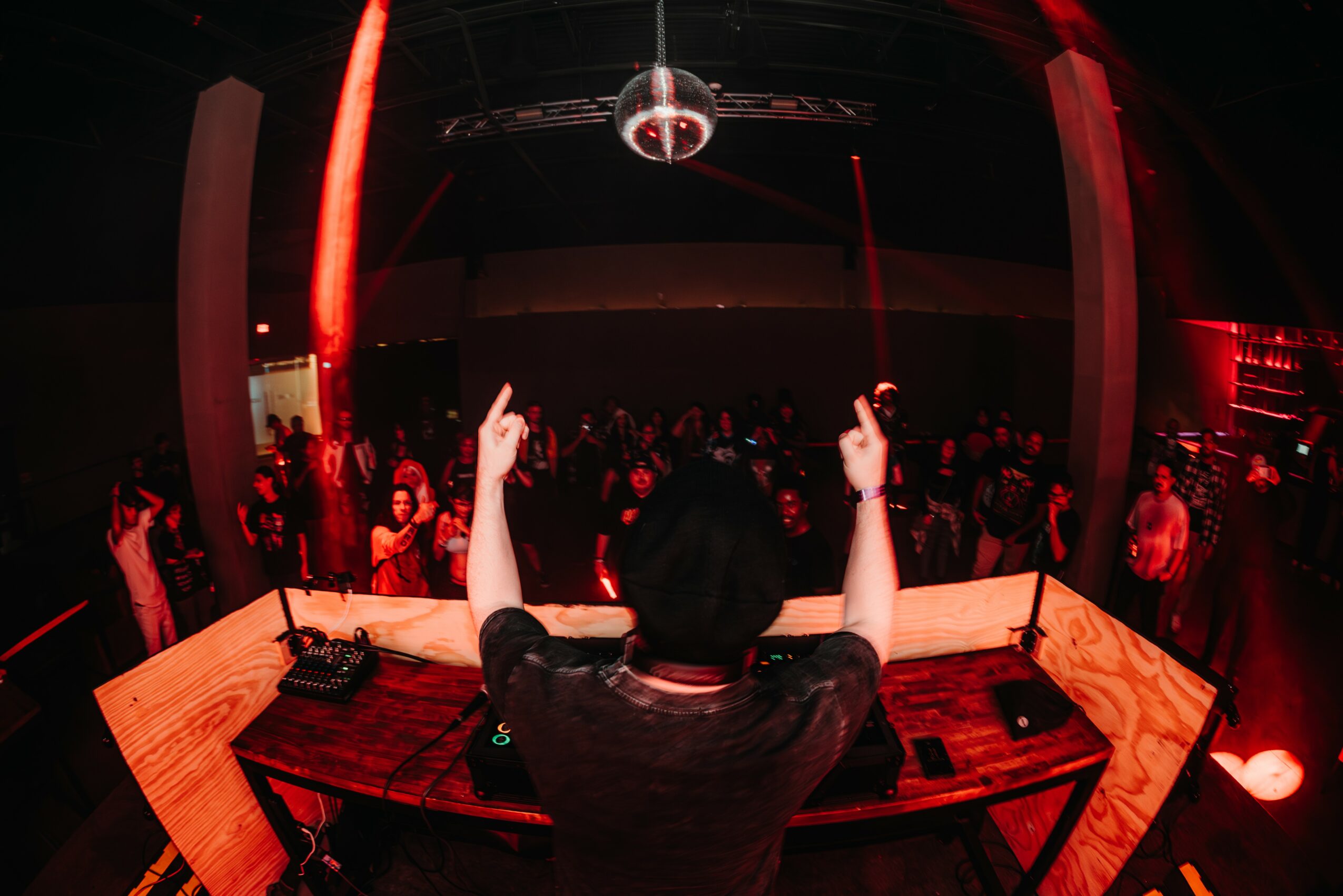 DJ with raised hands in front of a crowd at a nightclub, red lighting, disco ball overhead, wooden DJ booth, people dancing