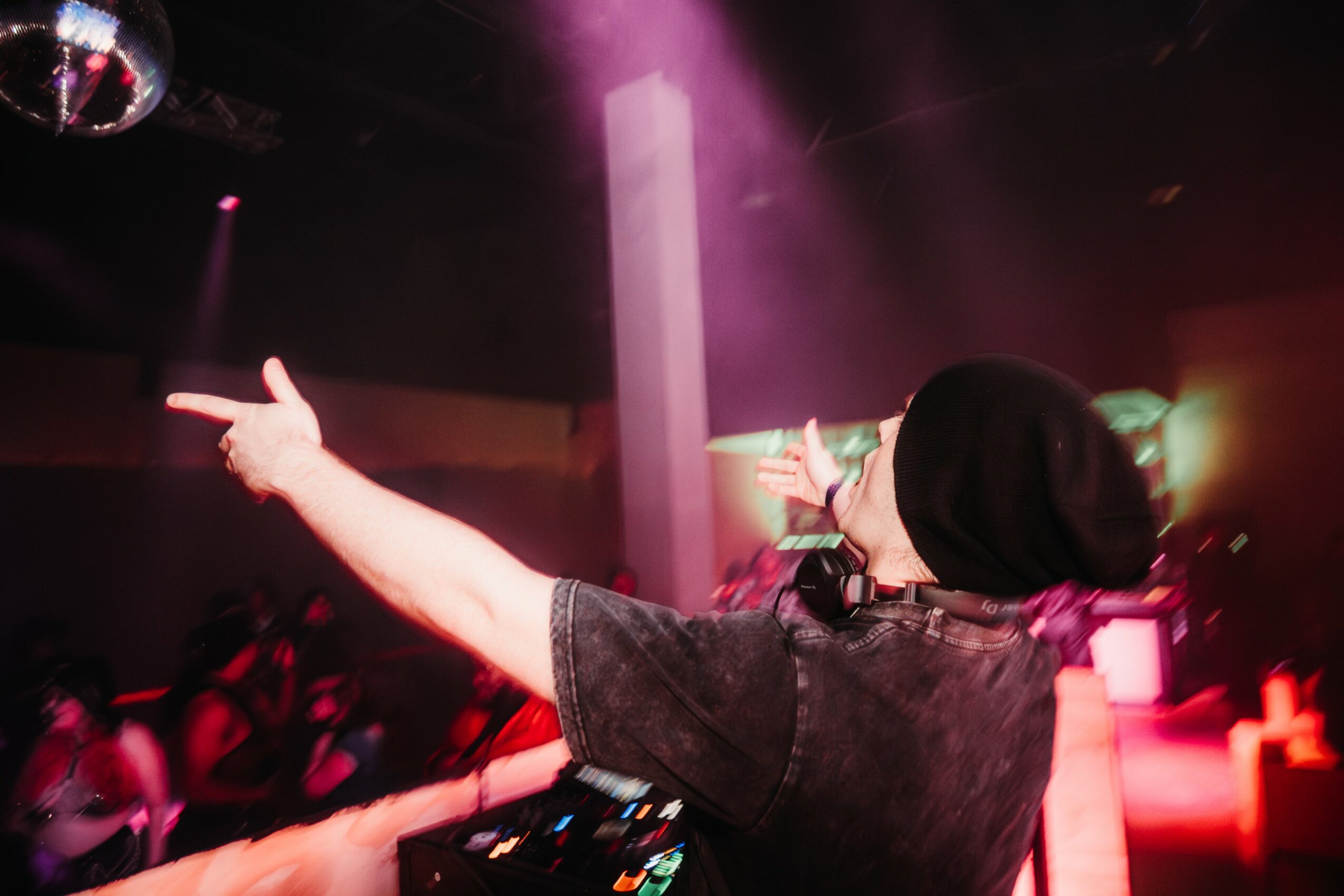 Person with black hair and dark clothing DJing at a club with pink and purple lighting, arms outstretched.