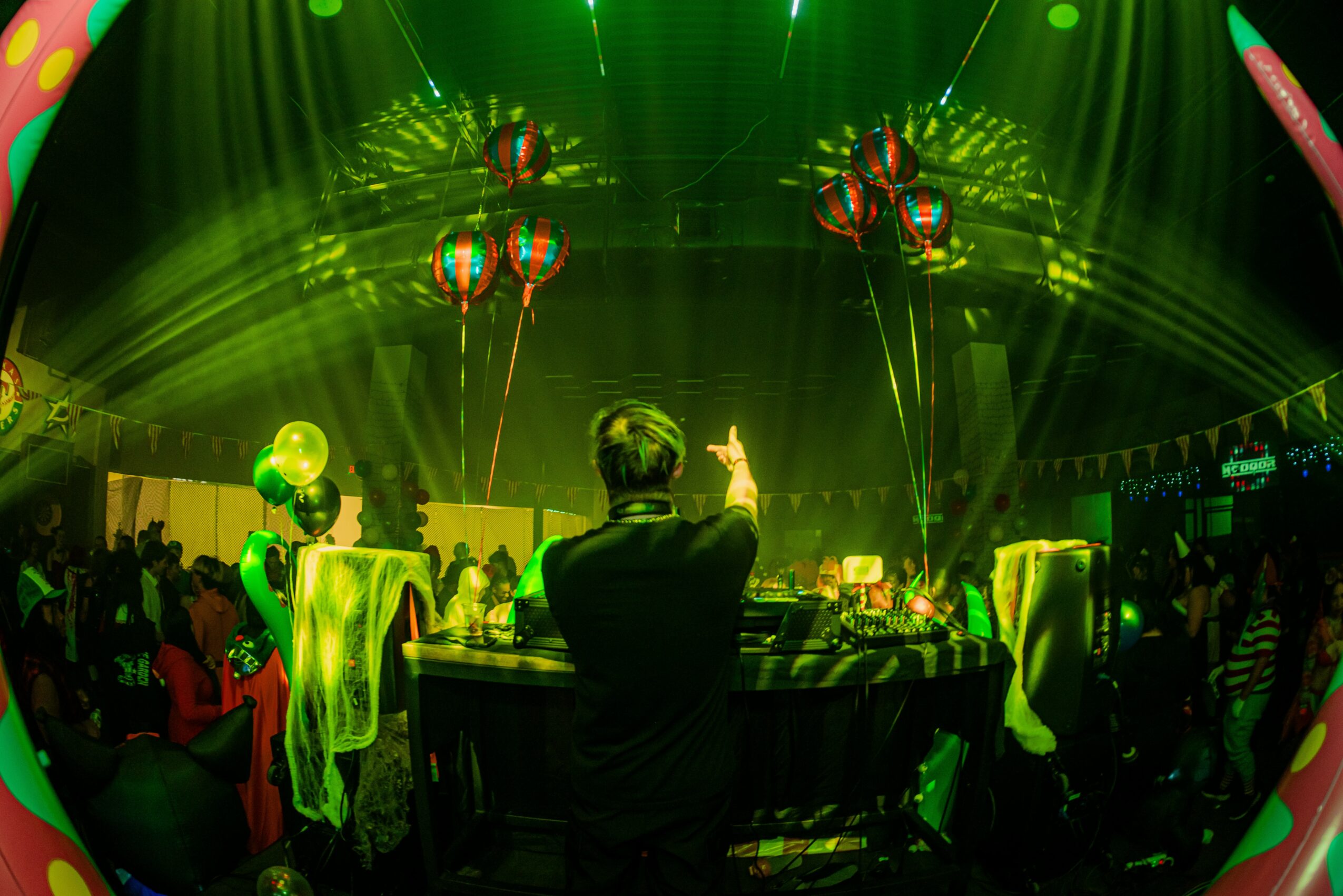 Person standing at DJ booth with colorful balloons and green laser lights in a lively party scene.