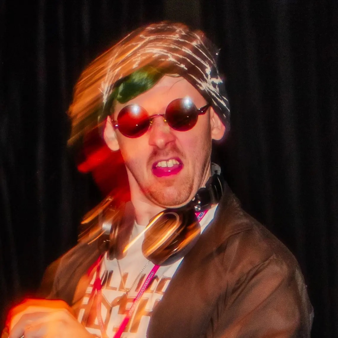 Person with sunglasses, headphones, and a headscarf, making a grimace, against a dark background.