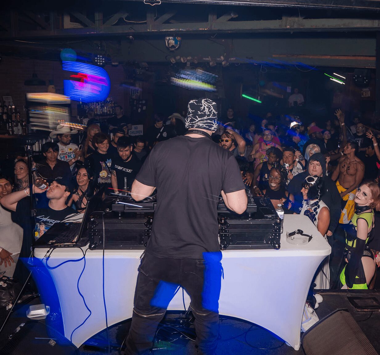 DJ wearing a patterned hat performs at a crowded nightclub with colorful lights and dancing people.