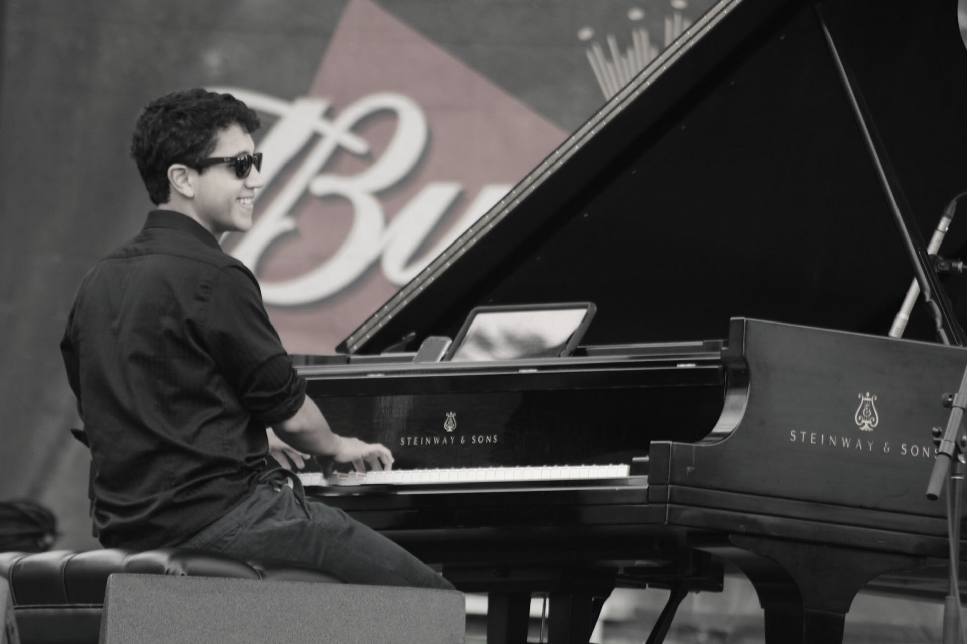Young person playing a grand piano outdoors, wearing sunglasses and a dark shirt, with a blurred background.